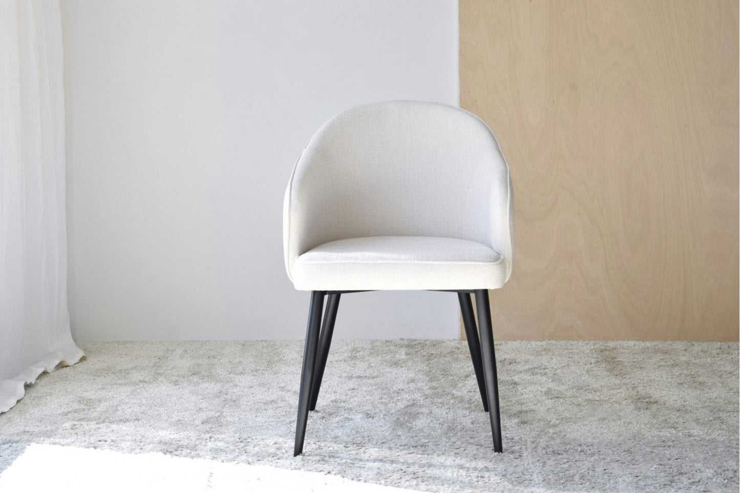 Una moderna silla beige del Pack 2 Sillas de Comedor con Reposabrazos Roma, con un diseño ergonómico y respaldo redondeado, se encuentra sobre una alfombra gris claro contra una pared complementaria blanca y beige, con elegantes patas negras.