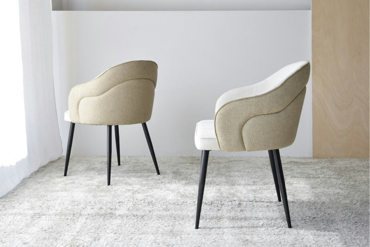 Dos sillas de comedor Roma con apoyabrazos y tapizado beige se encuentran sobre una alfombra gris texturizada en una habitación minimalista, con paredes neutrales y patas de metal negro.
