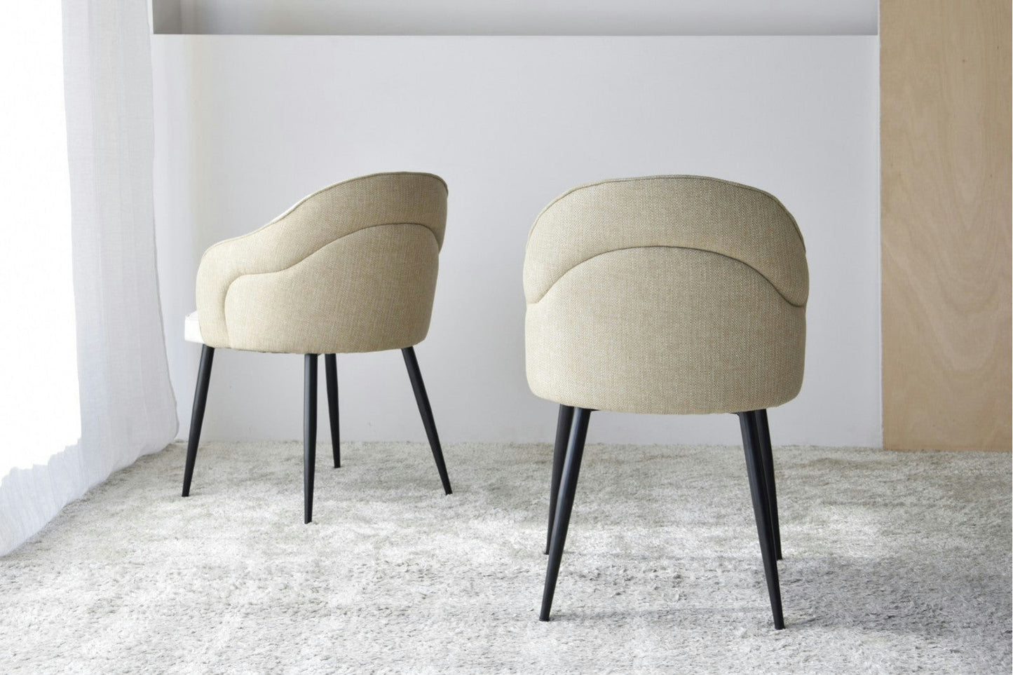 El Pack 2 Sillas de Comedor con Reposabrazos Roma (Beige) presenta tapizado beige y elegantes patas metálicas negras, elegantemente colocadas sobre una alfombra clara en una habitación minimalista.