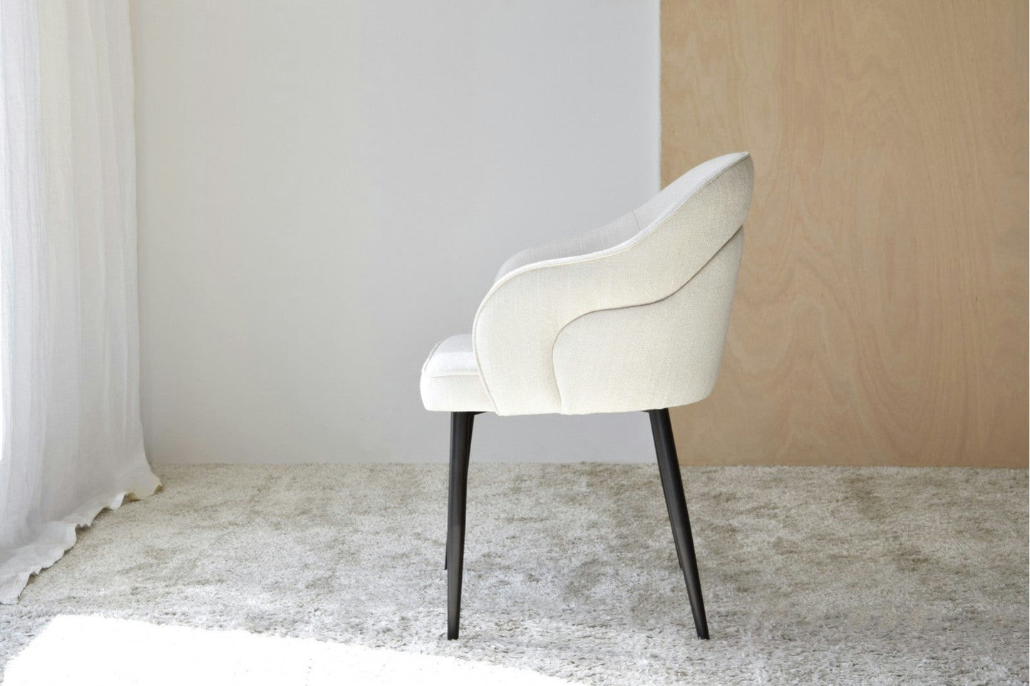 El Pack 2 Sillas de Comedor con Reposabrazos Roma en color beige claro, con patas negras, se encuentra sobre una alfombra clara. Colocado de lado contra una pared de dos tonos (mitad blanca y mitad con textura de madera), el conjunto se complementa con una cortina a la izquierda, que realza la elegancia moderna de este espacio.