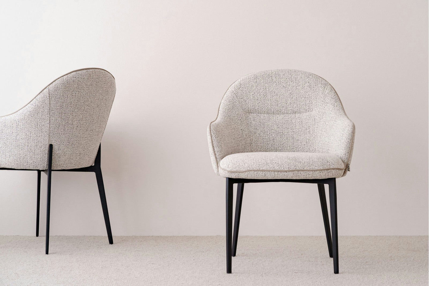 Dos sillas de comedor Elna en tonos beige y gris con respaldos curvos y patas negras, una orientada hacia adelante y la otra en ángulo. Esta disposición, que se apoya sobre un piso alfombrado en tonos beige y una pared en tonos beige, presenta un diseño elegante y contemporáneo.