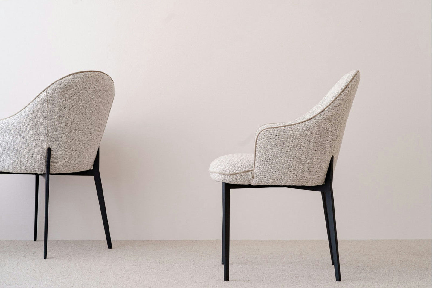 Dos sillas de comedor Elna en tonos beige y grises con patas negras se apoyan contra una pared blanca lisa, exudando un estilo moderno que combina a la perfección con su diseño contemporáneo.