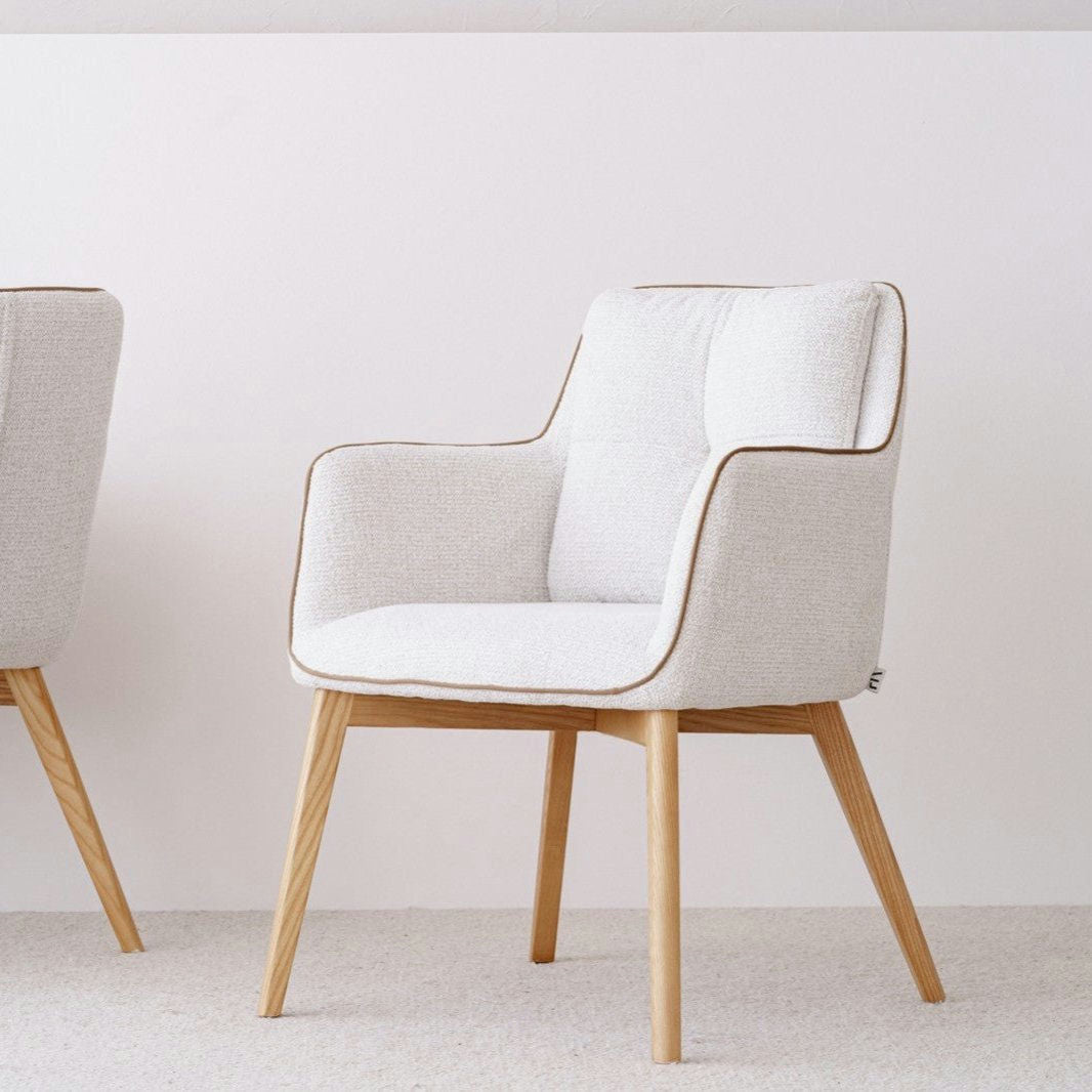 El Pack 2 Sillas de comedor tapizadas Marfil Oliver, tapizadas con poliéster, se alza elegantemente sobre una alfombra clara contra una pared blanca lisa. Una silla es parcialmente visible a la izquierda, agregando un sutil toque de estilo escandinavo con patas de madera.