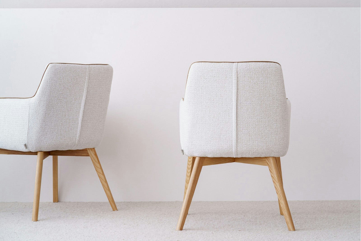 El Pack 2 Sillas de comedor tapizadas Marfil Oliver, con un diseño moderno y elegante con tapizado en gris claro y patas de madera, se coloca contra una pared blanca lisa sobre un piso alfombrado en color beige, encarnando el estilo escandinavo.