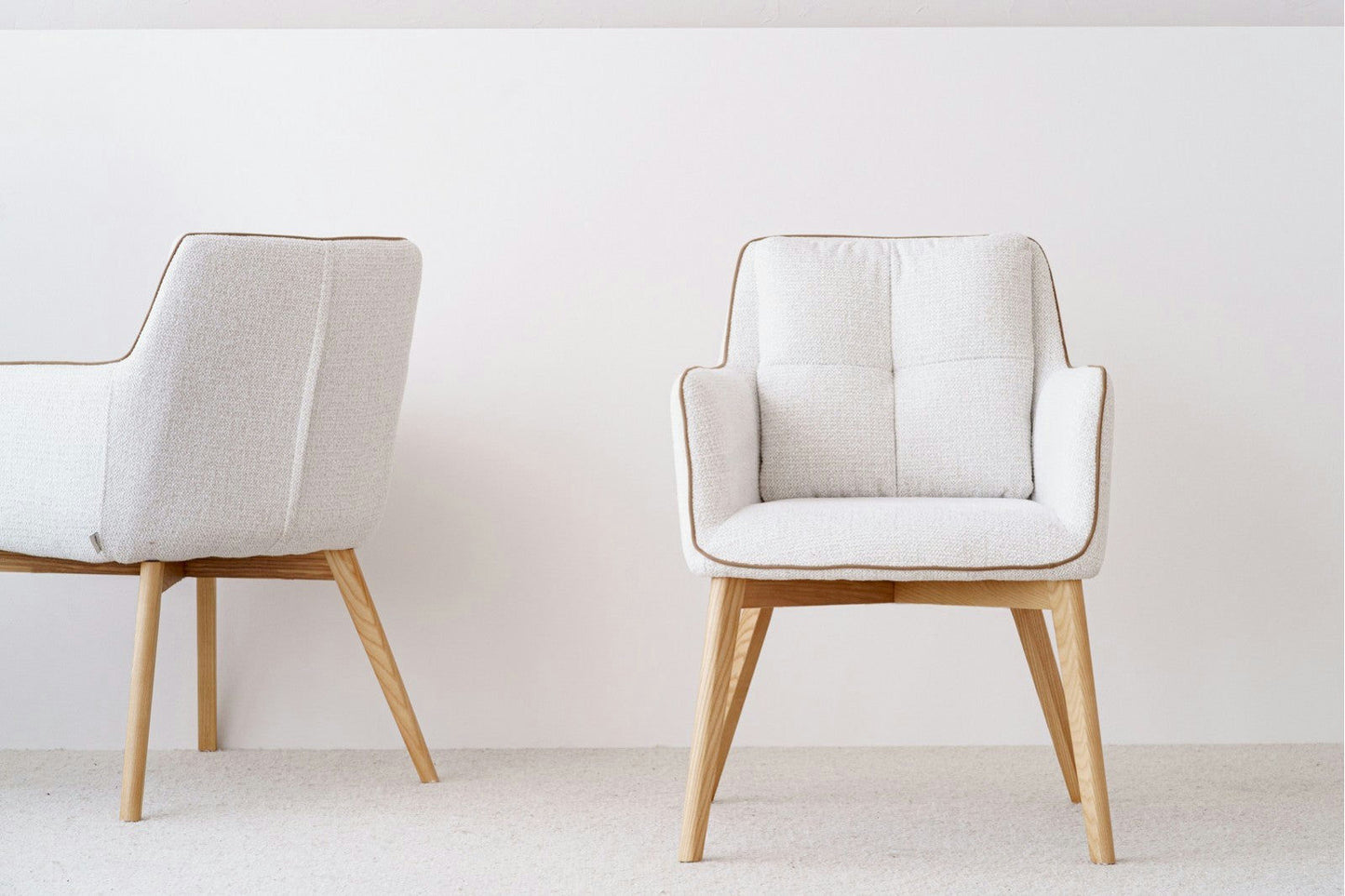 Dos elegantes sillas de comedor Marfil Oliver, con patas de madera, se encuentran sobre un piso alfombrado claro, exudando estilo escandinavo contra una pared blanca nítida.