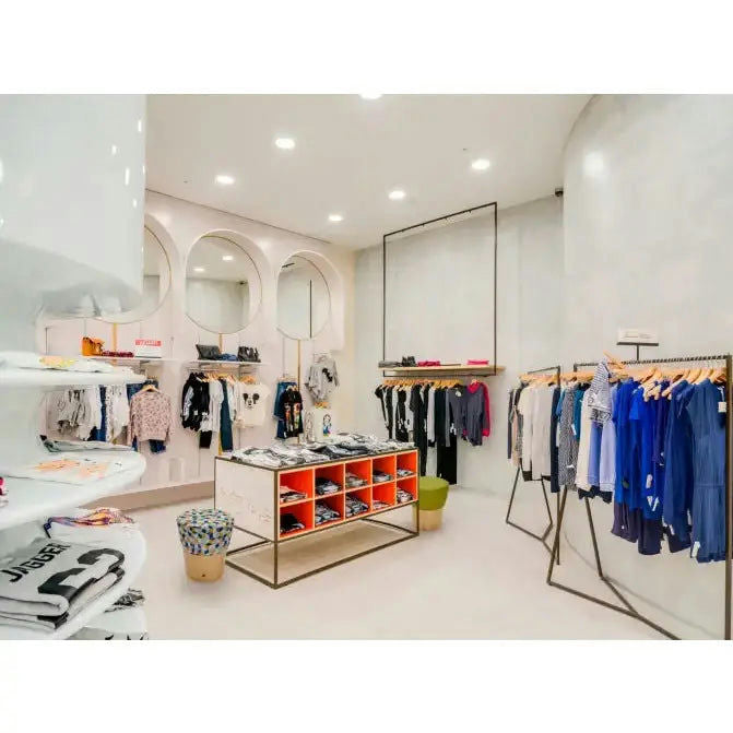 Interior de una tienda de ropa con percheros, una mesa de exposición central que muestra un Puff Redondo Tapizado y Madera de Pino Conny y grandes espejos. La decoración luminosa y minimalista cuenta con una estética escandinava limpia y moderna.