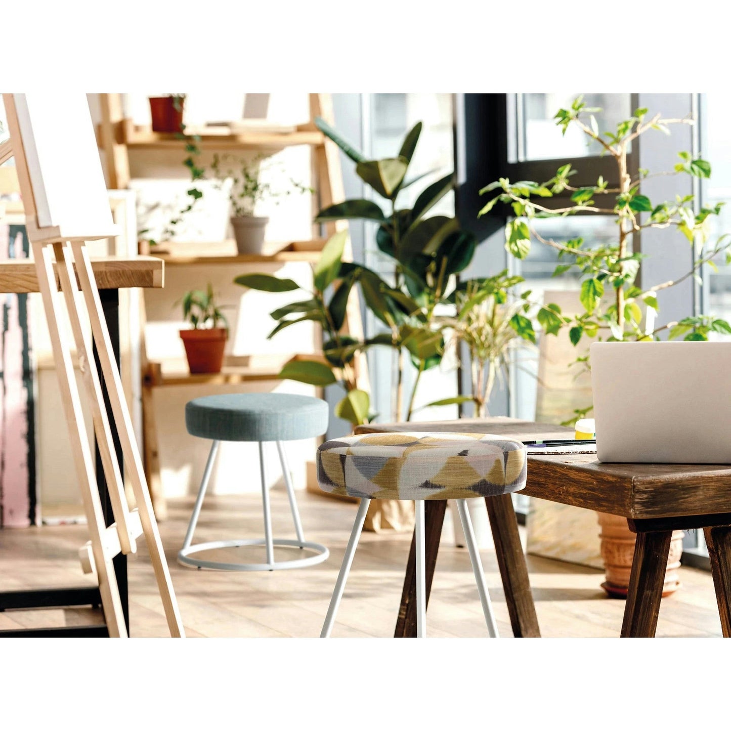 Una sala soleada cuenta con una mesa de madera, plantas, caballetes y dos taburetes. Una computadora portátil abierta reposa sobre la mesa, enmarcada por una elegante estructura Puff Redondo Tapizado y Metal Vita de diseño moderno. Los estantes del fondo están decorados con varias plantas en macetas.
