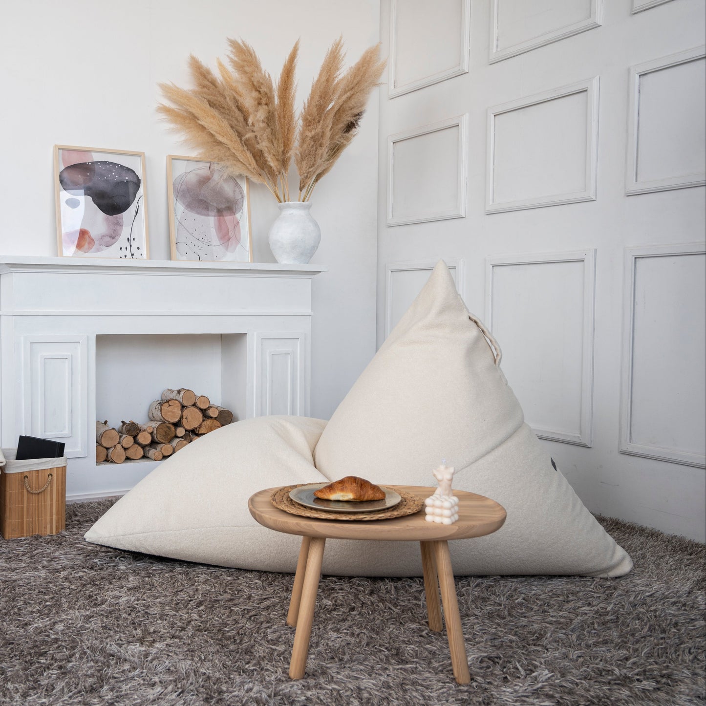 Una acogedora habitación con un Bean bag puff grande de tela borreguito 155×120×95 cm RAZZMATAZZ TEDDY, una mesita de madera con un cruasán y una decoración minimalista con arte abstracto y hierba de la pampa en un jarrón.