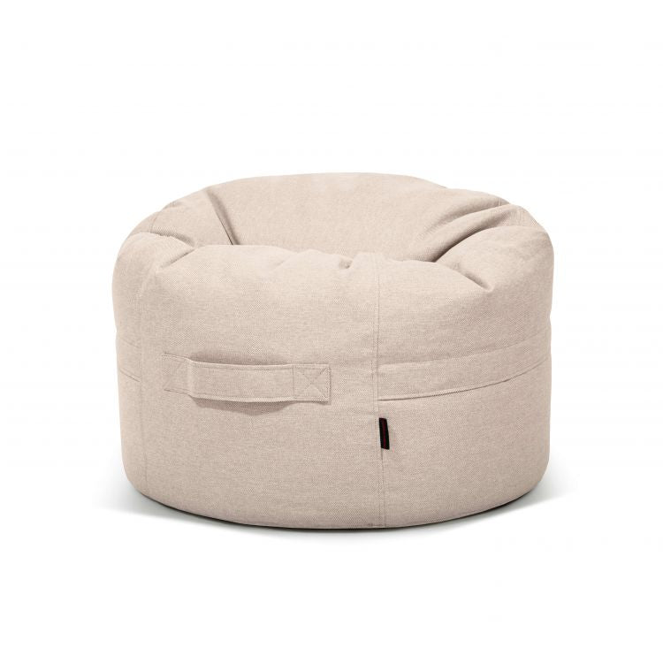 El ROLL 80 RIVIERA es un sillón bean bag redondo de tela beige (80×80×35 cm) con tejido hidrófugo y antimanchas, costuras visibles, asa lateral y etiqueta negra: versátil para uso en interiores y exteriores.