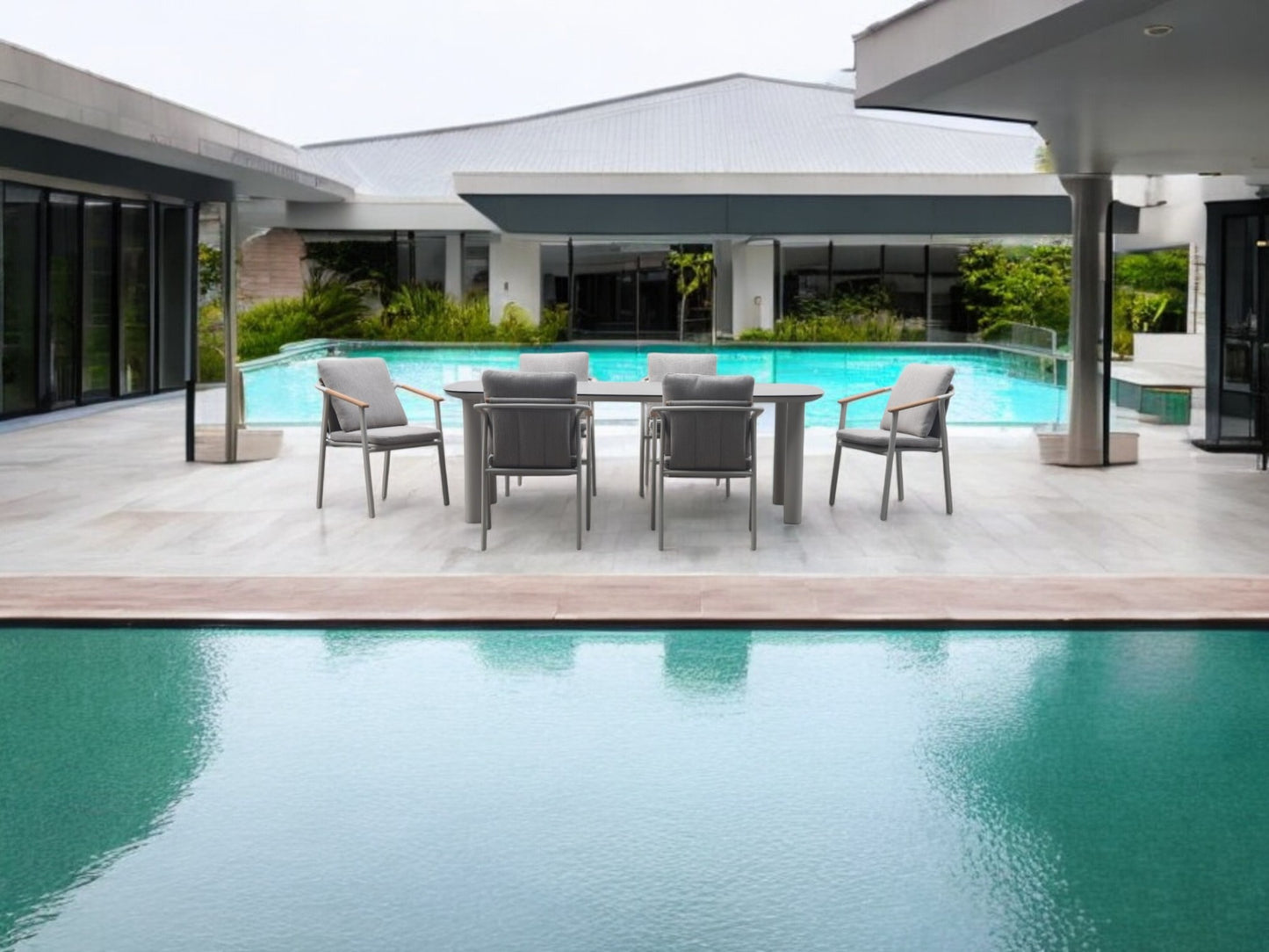 Una moderna mesa de comedor exterior con seis sillas y el Sillón de exterior en aluminio y madera de teca - gris arena - SAND se colocan en un patio de baldosas junto a una gran piscina, rodeado de edificios contemporáneos.