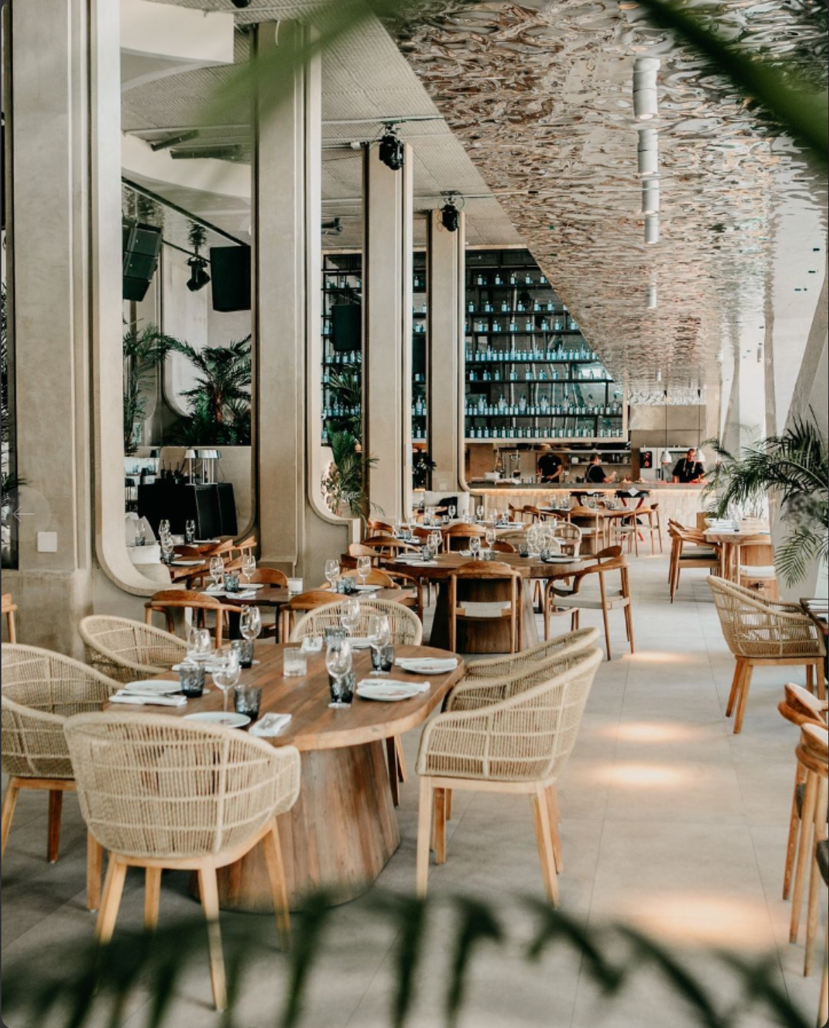 Un restaurante moderno y bien iluminado exhibe mesas de madera y sillas de mimbre Silla de comedor Ratán Teca Natural Corda debajo de un techo con patrones intrincados. El bar está completamente abastecido y las plantas junto con elementos de diseño artesanal realzan el encanto del espacio.