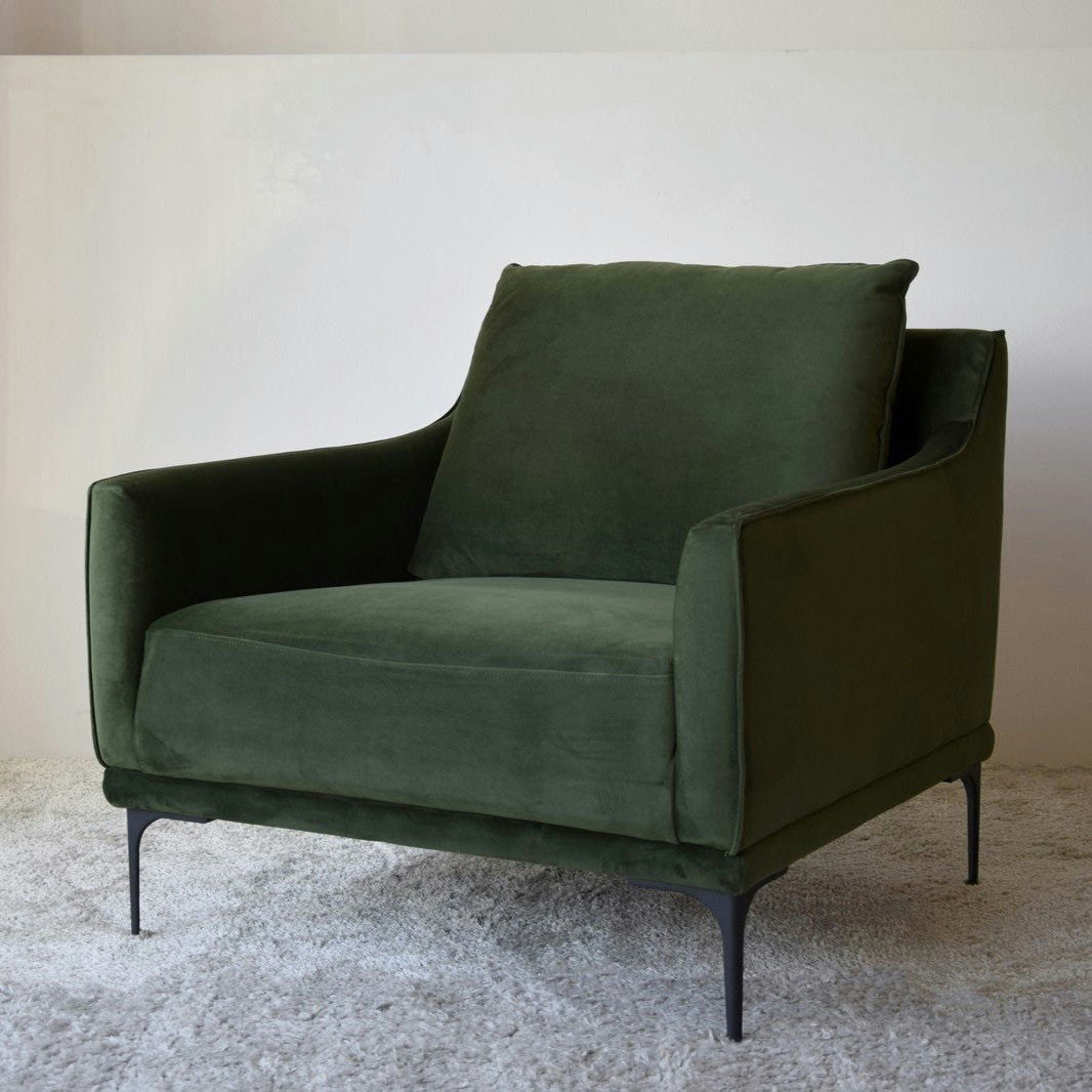 El Sillón de Diseño en Terciopelo Ntsu en terciopelo verde oscuro tiene un asiento y respaldo acolchados, que descansan elegantemente sobre patas de metal sobre una alfombra blanca impecable.