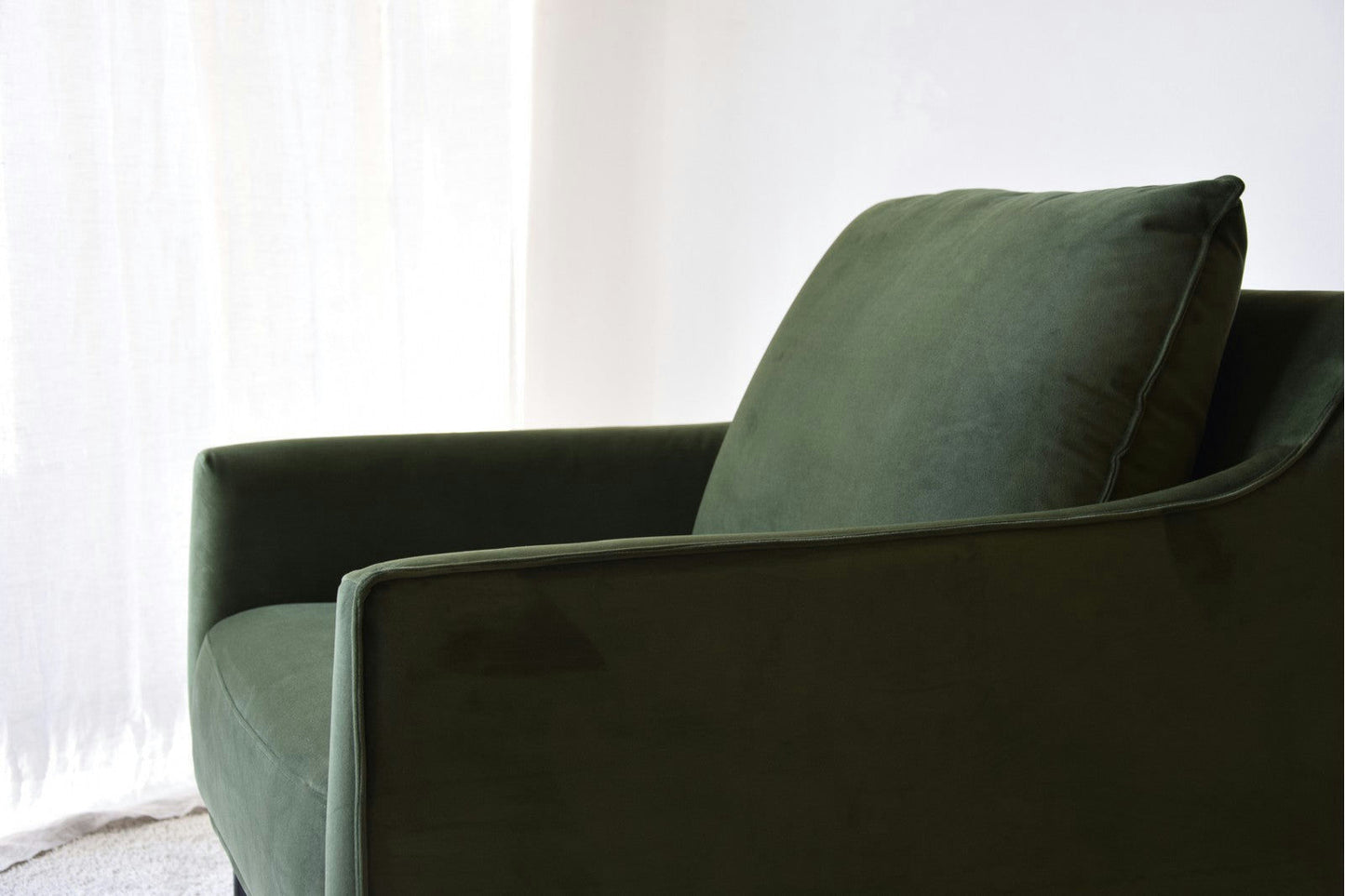 Un sillón de Diseño en Terciopelo Ntsu de terciopelo verde oscuro con elegantes patas de metal y un cojín, colocado en una habitación bien iluminada junto a una cortina blanca, realza cualquier espacio con sofisticación y estilo.