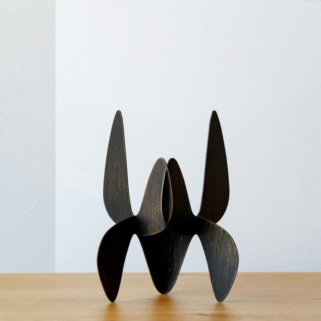 Una moderna escultura abstracta negra con formas curvas, elaborada en metal orgánico, se exhibe con estilo sobre una superficie de madera contra una pared blanca sencilla.