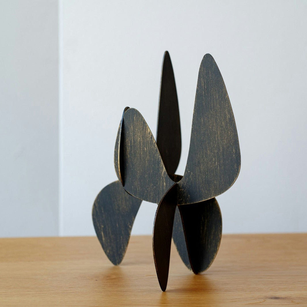 Una llamativa escultura negra con formas curvas y superpuestas se exhibe sobre madera. Ubicada sobre un fondo claro y sencillo, esta cautivadora pieza se integra en su entorno de manera muy similar a las Figuras Decorativas en Metal Orgánico Lámina Oro.