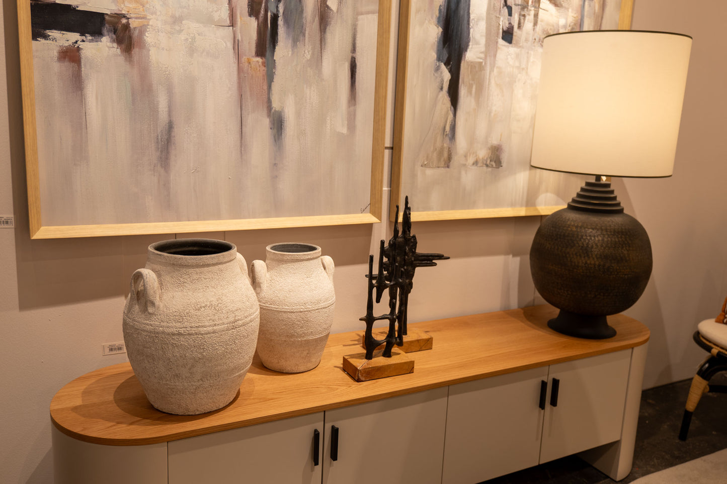Un aparador de madera exhibe dos jarrones de cerámica blanca, una pieza escultórica negra y la Lámpara de mesa en Metal N.9 Aurum, con cuadros abstractos colgados encima.