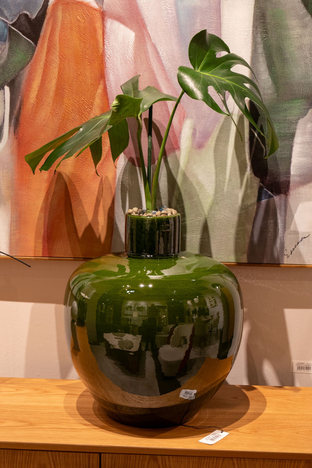Un gran jarrón verde brillante de la Colección Jarrones en Cerámica. Colección Arume con acabado verde musgo y una planta monstera sobre una superficie de madera ante un colorido cuadro abstracto.
