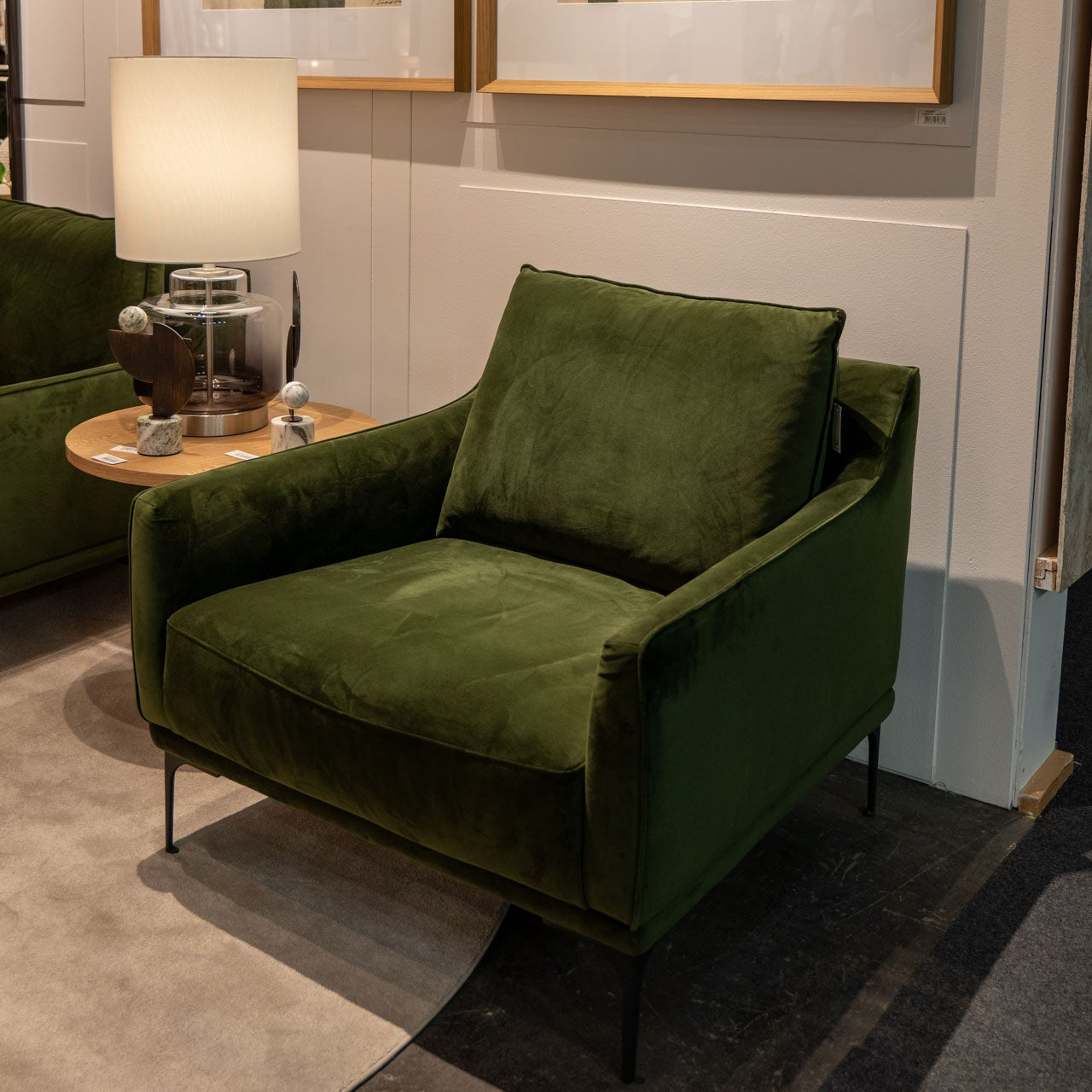 El Sillón de Diseño en Terciopelo Ntsu, en verde oscuro con patas negras, se ubica junto a una mesa auxiliar redonda de madera con lámpara y decoraciones, sobre una alfombra clara en una sala moderna.