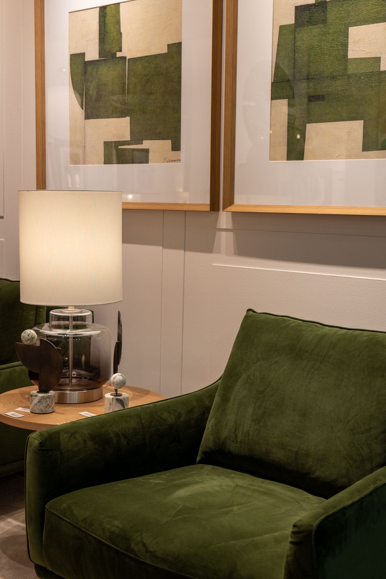 El Sillón de Diseño en Terciopelo Ntsu en verde oscuro se sitúa junto a una mesa auxiliar de madera con lámpara y objetos decorativos, bajo cuadros abstractos que adornan la pared.