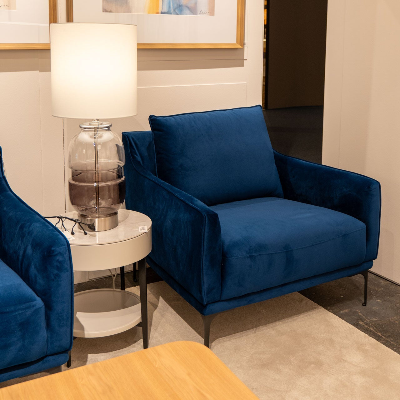 La butaca Helia en terciopelo azul noche se encuentra junto a una mesa auxiliar redonda con una lámpara de cristal y unas gafas, todo sobre una alfombra beige.