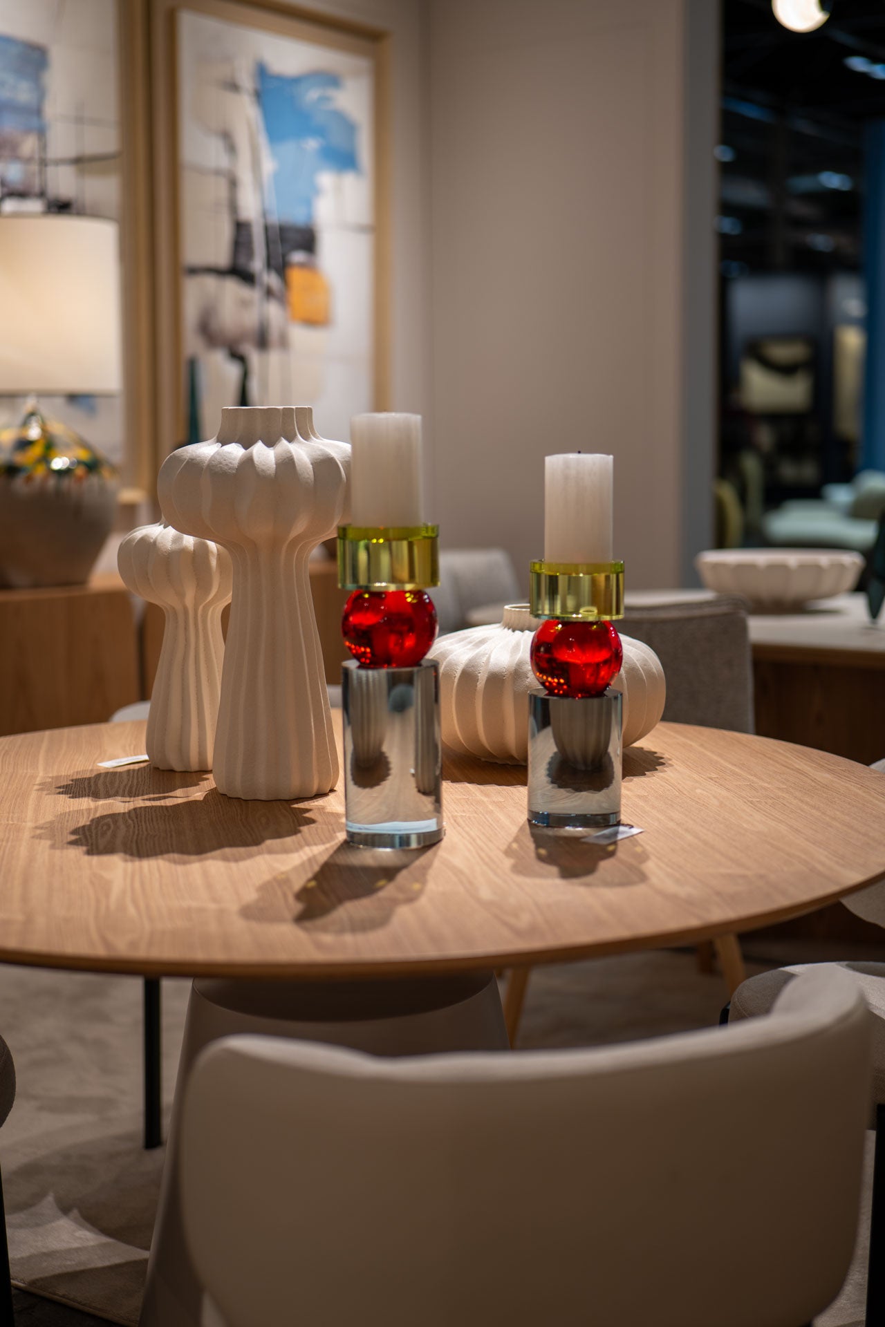 Una mesa redonda de madera con Candelabros Cristal Color Cilindro (grande y pequeño), jarrones de cerámica blanca y dos portavelas de cristal con detalles en rojo y dorado, rodeada de sillas modernas, ideal para una decoración elegante del hogar.