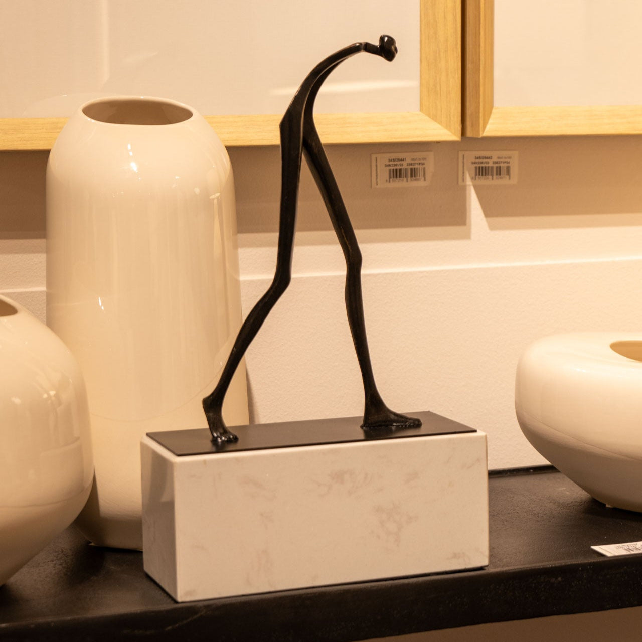 La Escultura Caminante, con una esbelta figura andante de metal negro sobre una base de mármol blanco, se alza entre grandes jarrones de cerámica blanca, creando un llamativo contraste decorativo.