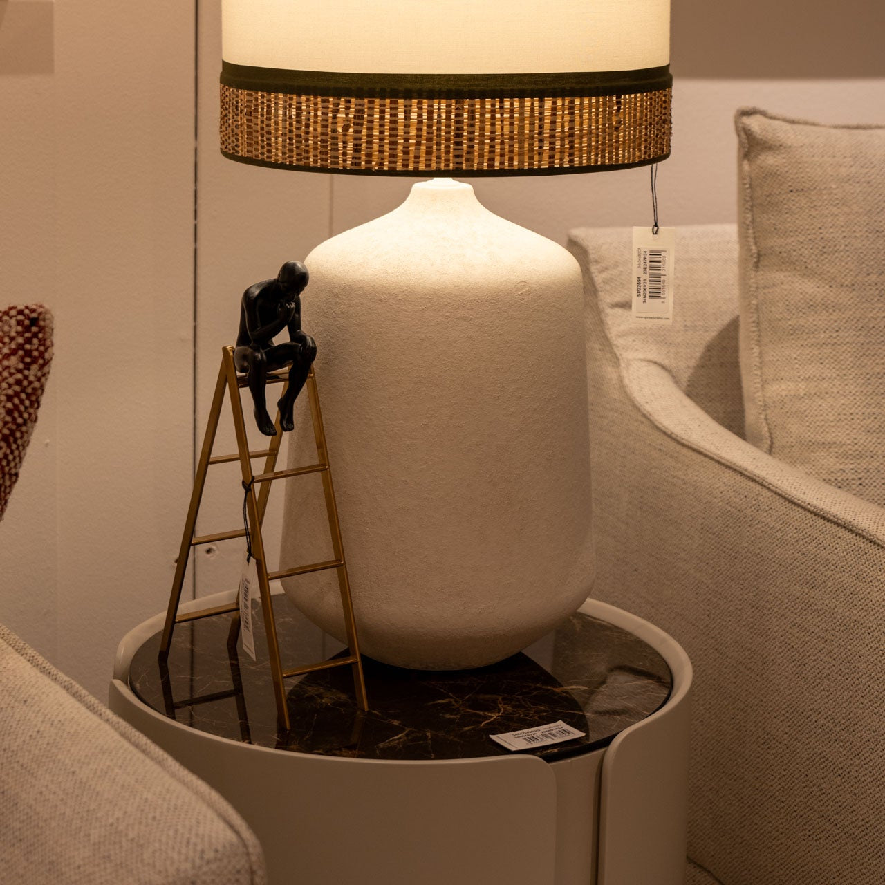 Una Lámpara de mesa en Cerámica Cíes con un ribete tejido descansa sobre una mesa auxiliar redonda; junto a ella, una pequeña escultura negra de una figura subiendo una escalera se apoya en la lámpara.