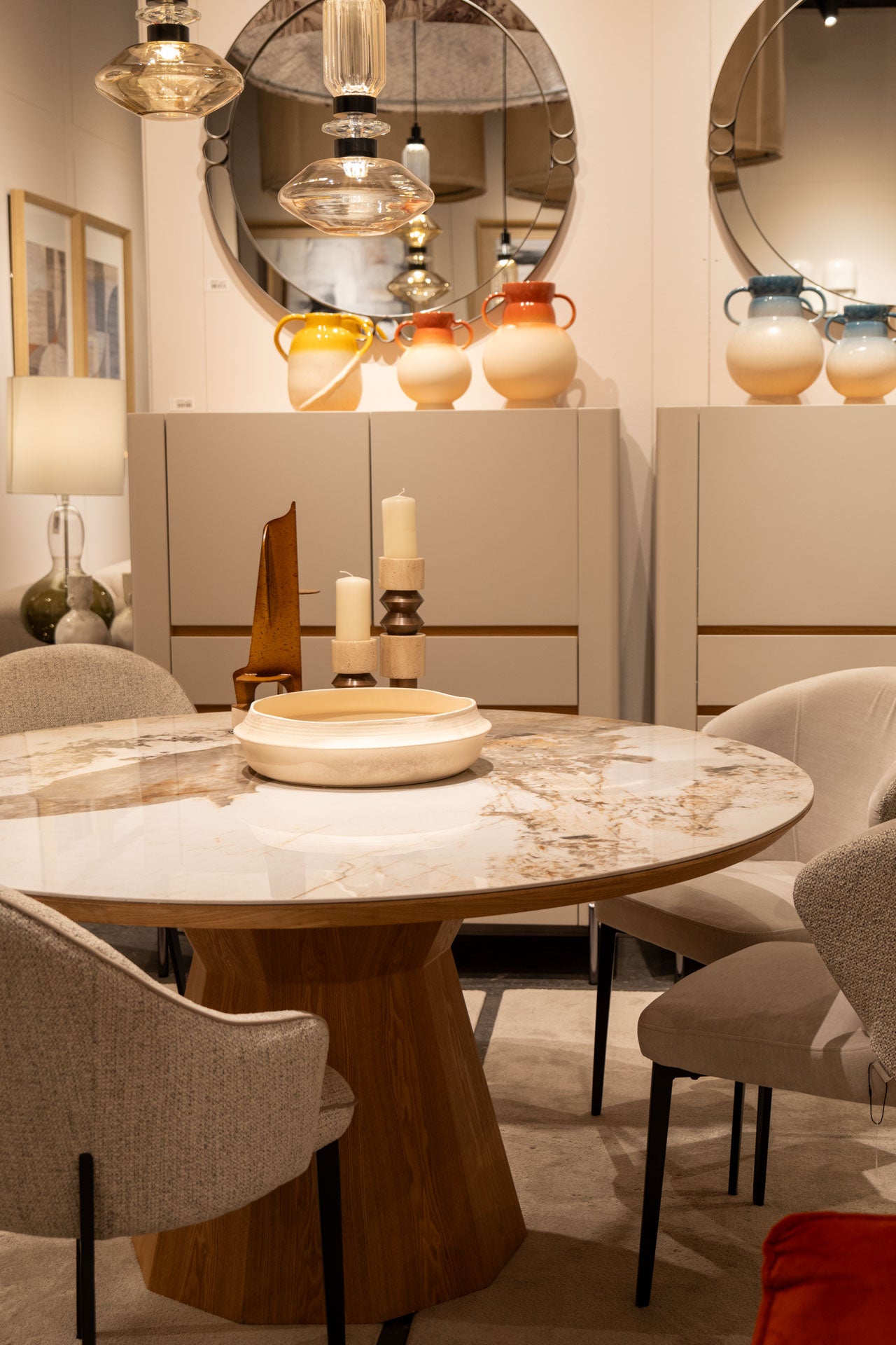 Un comedor moderno con la Mesa de comedor redonda en cerámico y fresno Nat Mistral, cuatro sillas tapizadas, velas decorativas, lámparas colgantes, armarios con jarrones de colores y grandes espejos redondos.