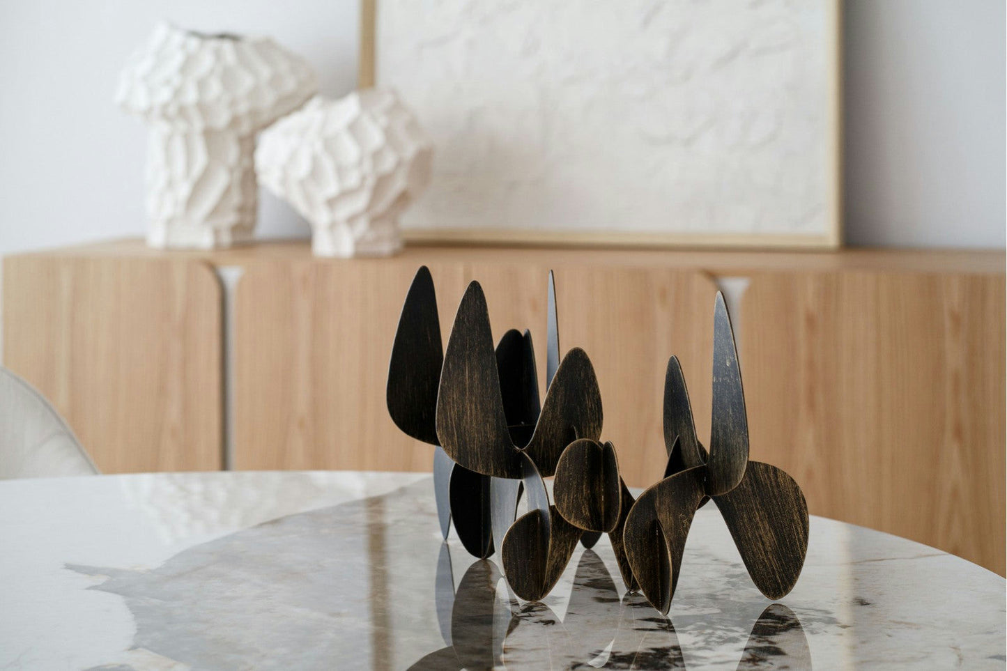 Escultura abstracta de color negro de la colección Figuras Decorativas en Metal Orgánico Lámina Oro sobre mesa de mármol, con lámparas blancas texturizadas y muebles de madera al fondo.