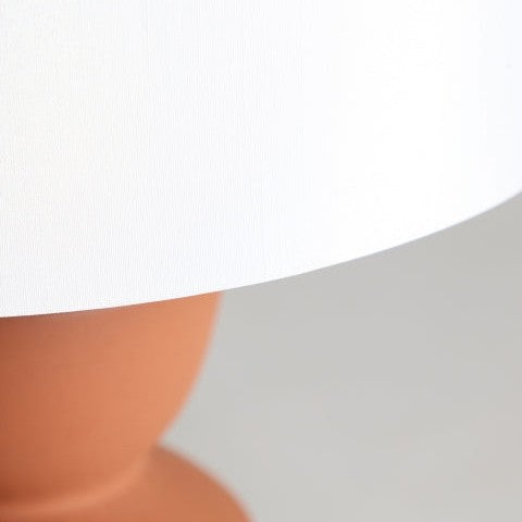 Primer plano de una pantalla blanca sobre la base redonda de color terracota de la Lámpara de mesa en cerámica Hasna sobre un fondo neutro, ideal para añadir iluminación suave y un toque de diseño atemporal al espacio.