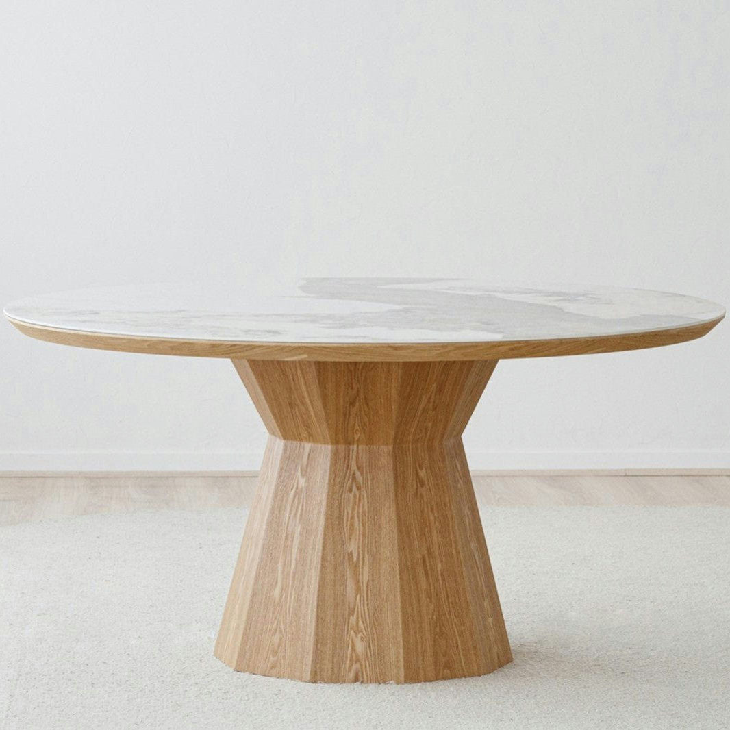 La Mesa de comedor redonda en cerámico y fresno Nat Dread se alza sobre una alfombra de colores claros en una habitación minimalistamente decorada.