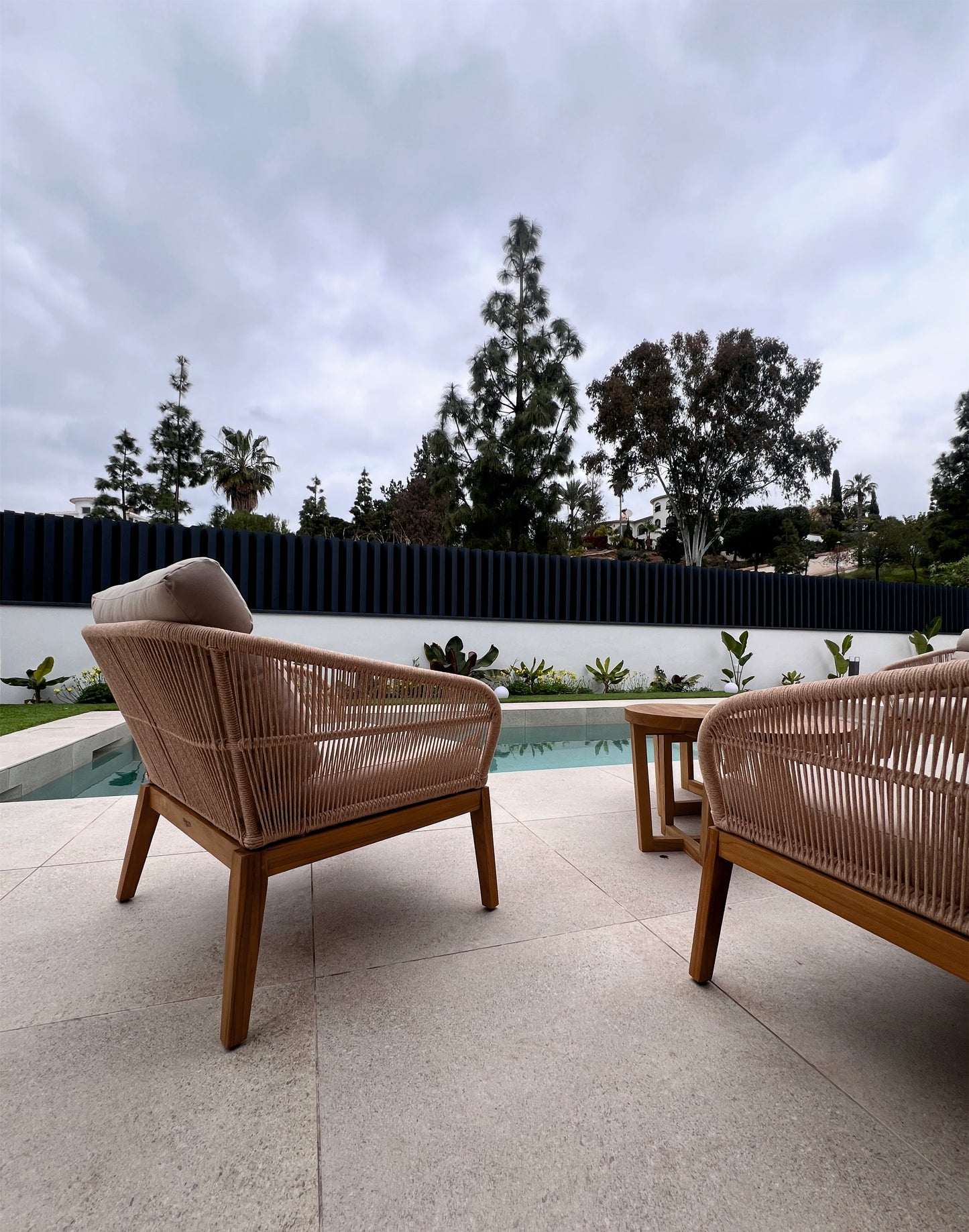 Dos sillas de mimbre y una mesita se sientan en un patio de piedra junto a la piscina, acentuado por el Sillón de exterior de teca y cuerda sintética beige 84 × 77 × 75 cm - natural - NATURE, rodeado de árboles-ideal para los amantes de la decoración de jardines.