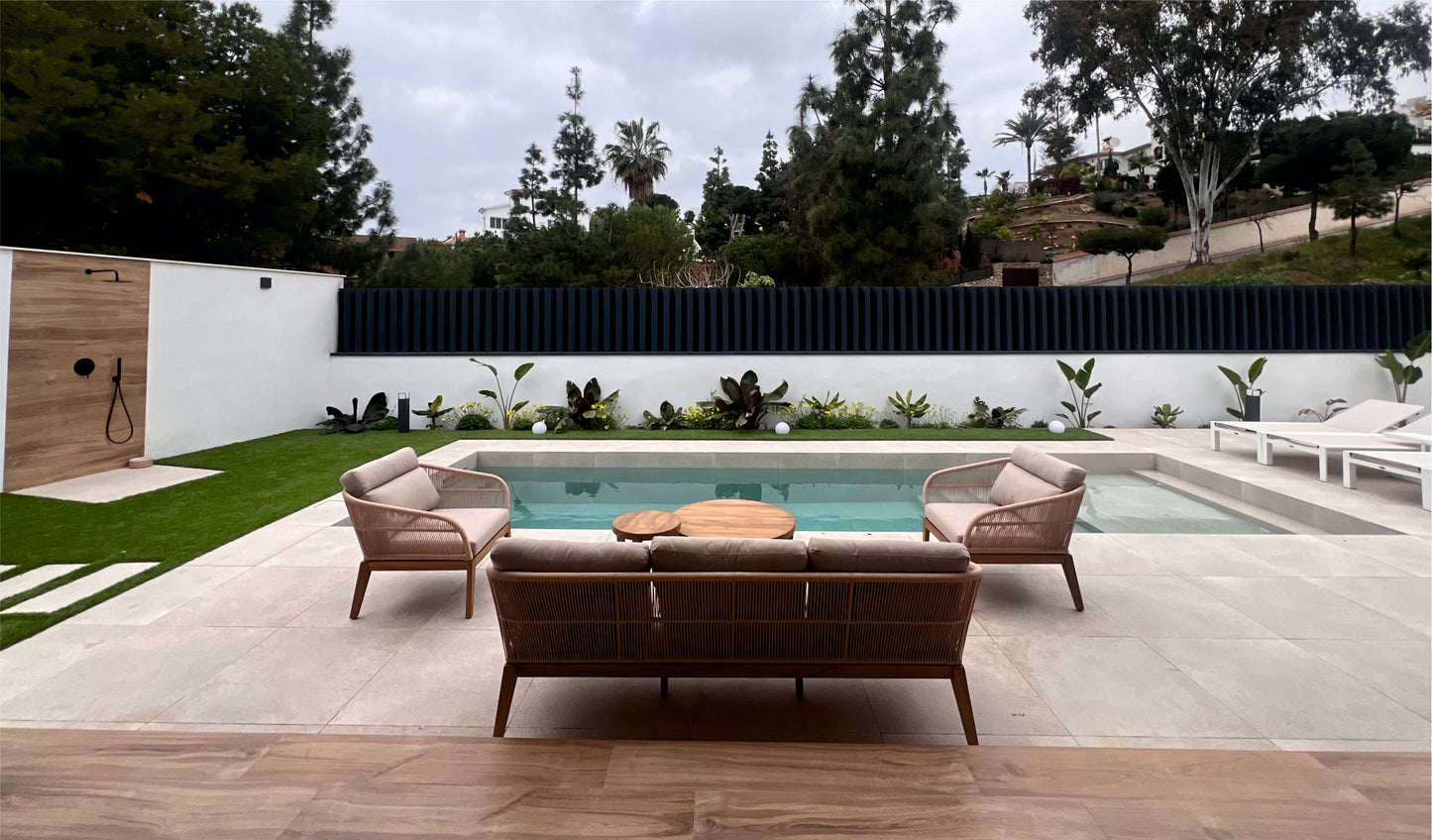 El moderno patio trasero cuenta con una pequeña piscina, dos tumbonas, un sillón de exterior NATURE de teca y cuerda sintética beige (84×77×75 cm), una ducha exterior y una exuberante vegetación bajo un cielo nublado.