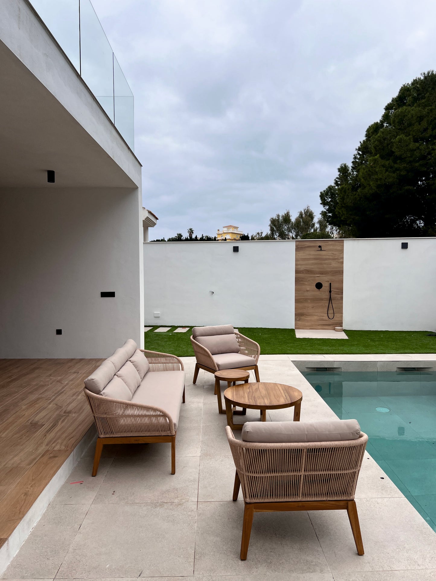 Moderno patio exterior con un sofá NATURE de 3 plazas de cuerda sintética beige y teca (200×77×75 cm), mesas de madera, junto a una piscina, delimitada por paredes blancas y una zona de césped con una ducha.