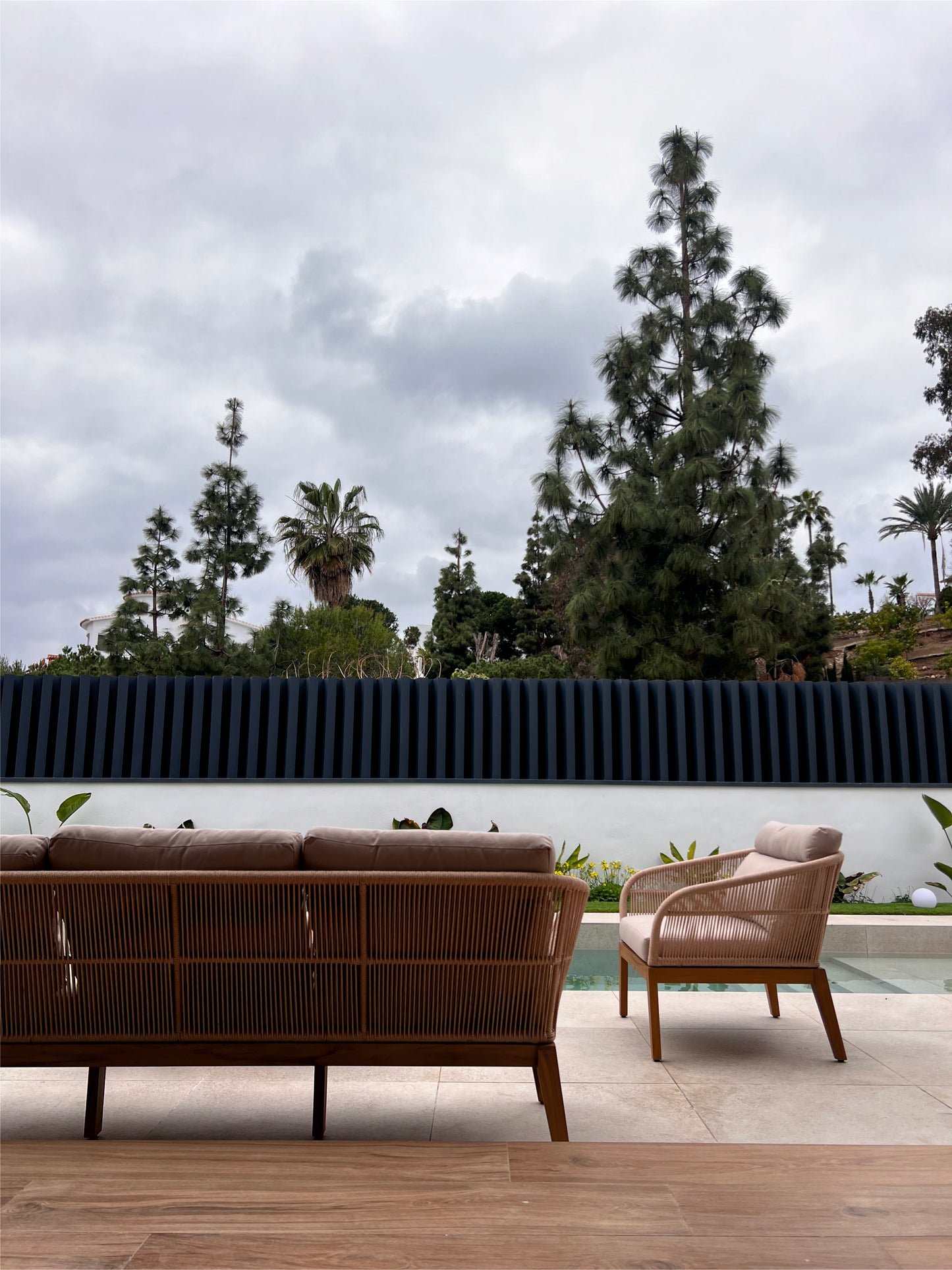 Un par de sillas Silla de exterior de teca y cuerda sintética beige NATURE se sientan en un patio embaldosado junto a una pequeña piscina, enmarcada por altos árboles y un cielo nublado.