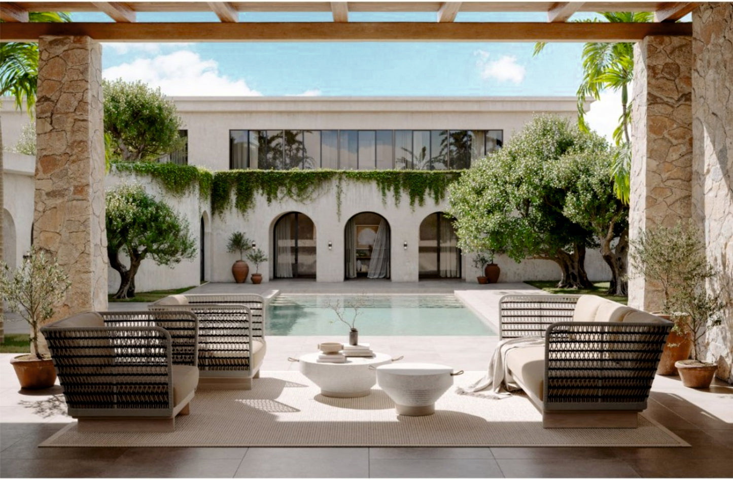 Un lujoso patio con modernos muebles de exterior de mimbre da a una piscina rectangular, enmarcada por columnas de piedra, plantas en macetas y una villa de dos plantas con arcos cubiertos de hiedra y una elegante mesa de centro redonda de teca NIZA (Ø100 × 25 cm) para exterior.