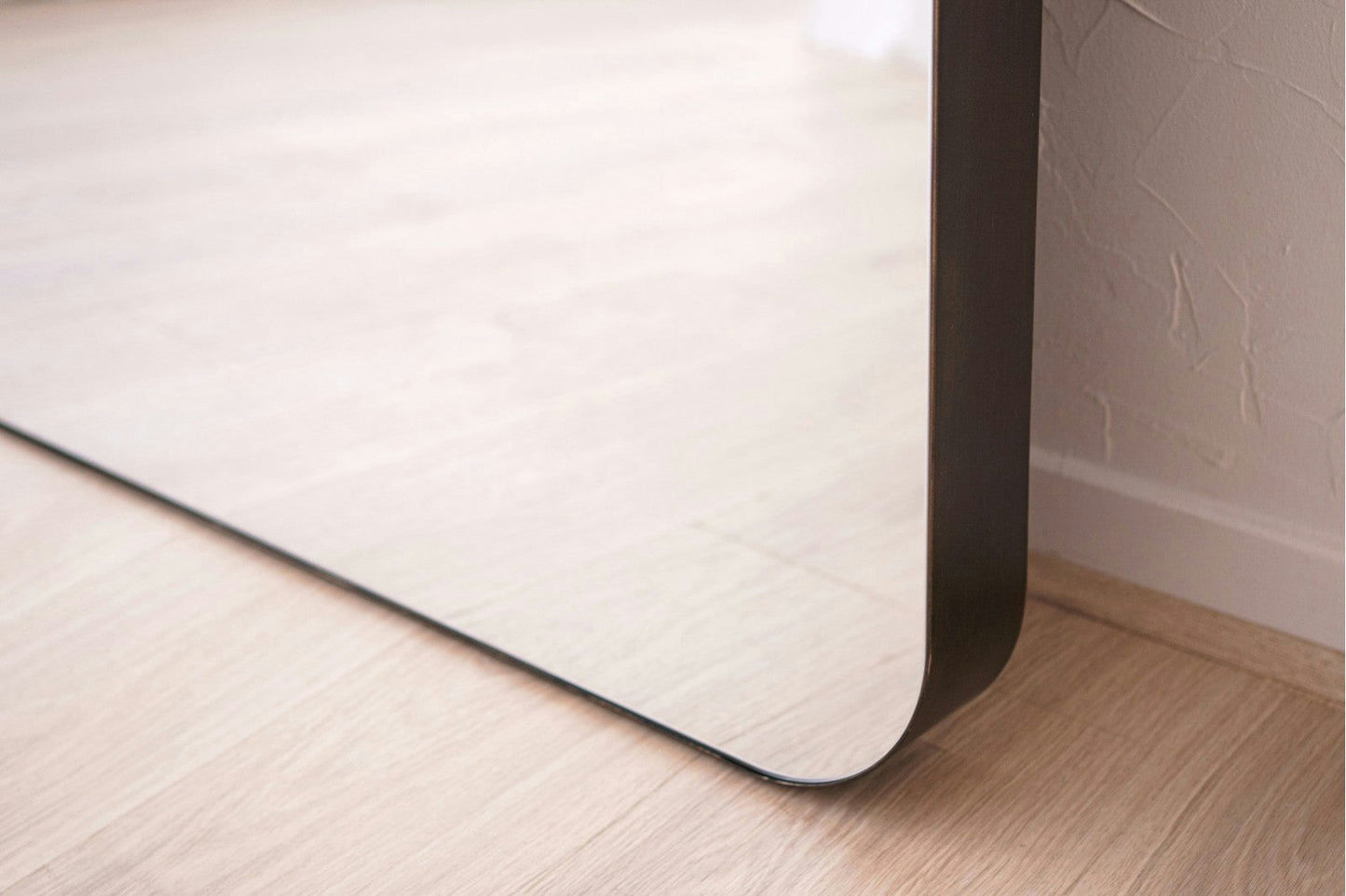 Primer plano de la esquina inferior del Espejo de suelo en metal industrial N4, con bordes redondeados y un marco de metal, apoyado contra una pared blanca texturizada sobre un piso de madera clara, perfecto para agregar estilo industrial.