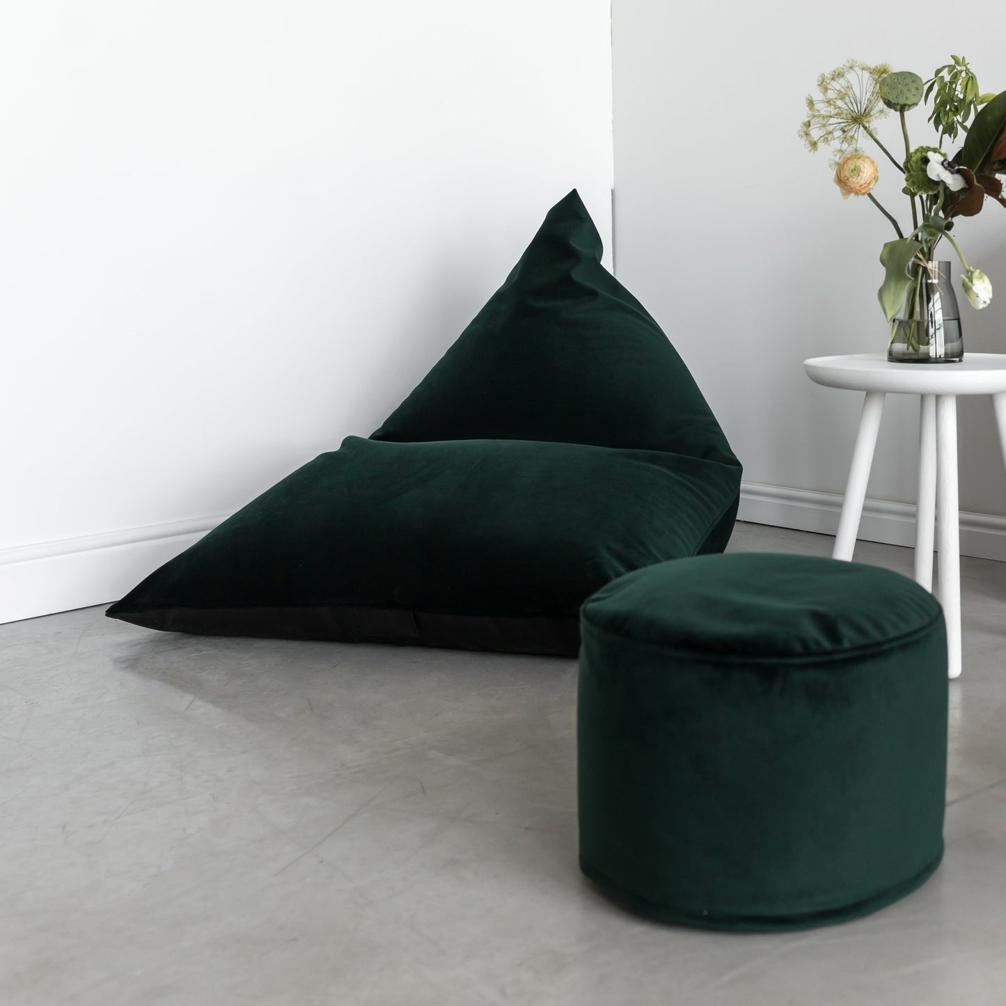 El Bean bag puff juvenil de terciopelo velour 110×130×90 cm by RAZZ BARCELONA y una otomana a juego en verde oscuro descansan sobre un suelo gris junto a una mesa blanca con un jarrón de cristal con flores contra una pared blanca.