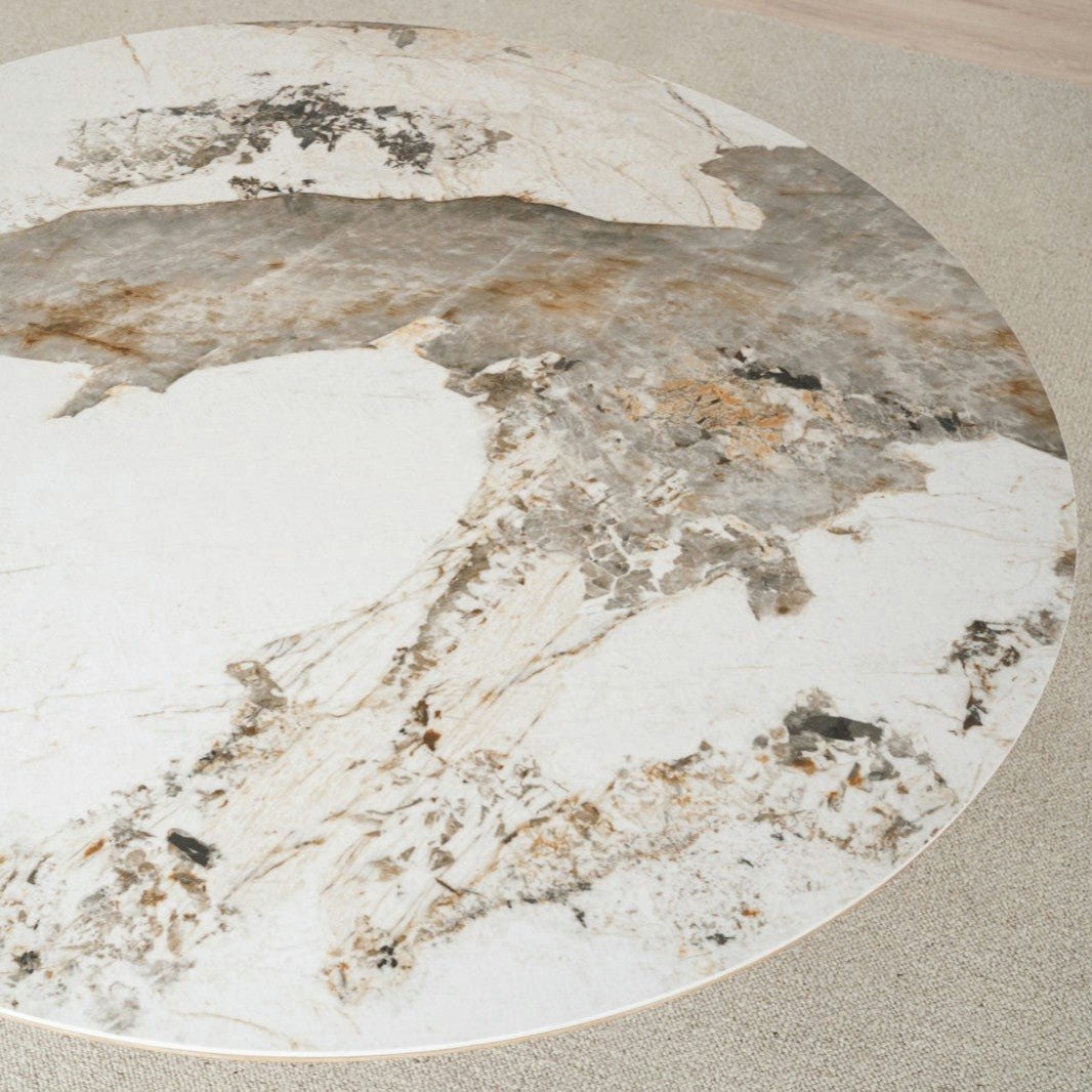 La mesa de comedor redonda de cerámica y fresno Nat Dread, con una superficie de mármol y cerámica que muestra patrones en blanco, gris y marrón, reposa elegantemente sobre un piso alfombrado de color claro. Su base captura el encanto natural de la madera fresca de Fresno.