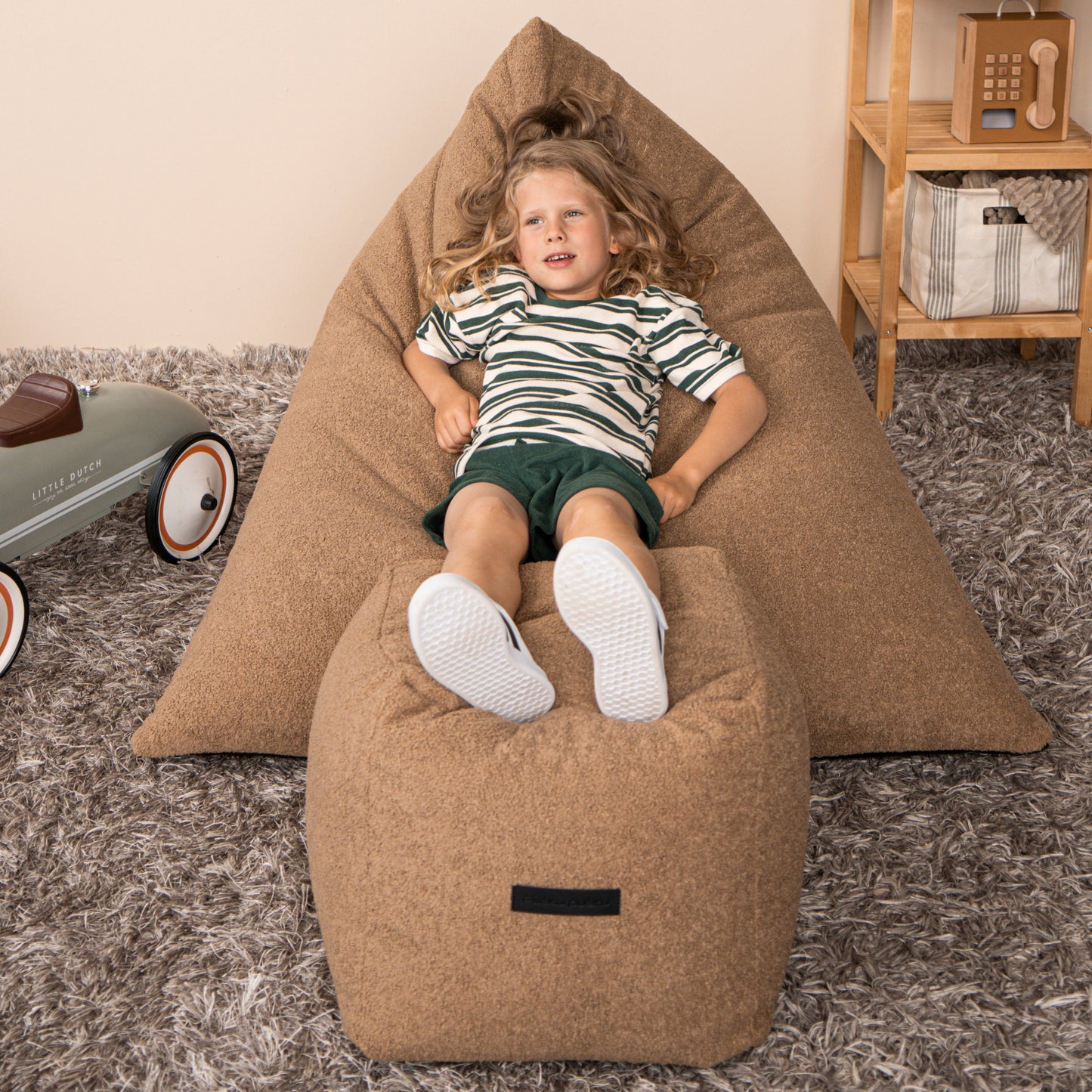 Un niño de pelo largo y rubio se relaja en un puff Bean bag juvenil de tela borreguito 110×130×90 cm -moderno- dormitorio - RAZZ TEDDY, con los pies en alto sobre una otomana a juego en una habitación alfombrada y llena de juguetes.