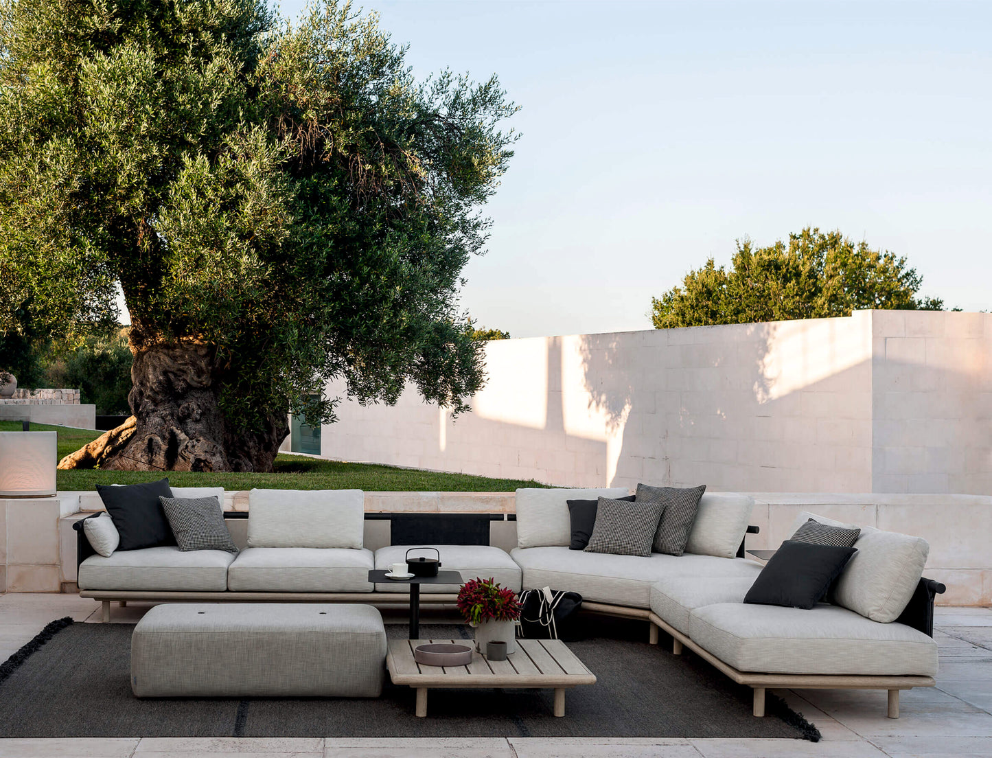 Moderno patio exterior con un sofá seccional beige, cojines grises, una teca SAMBA mesa de centro de exterior cuadrada (90×90×30 cm), y un gran olivo situado contra un muro de piedra blanca y un cielo despejado.