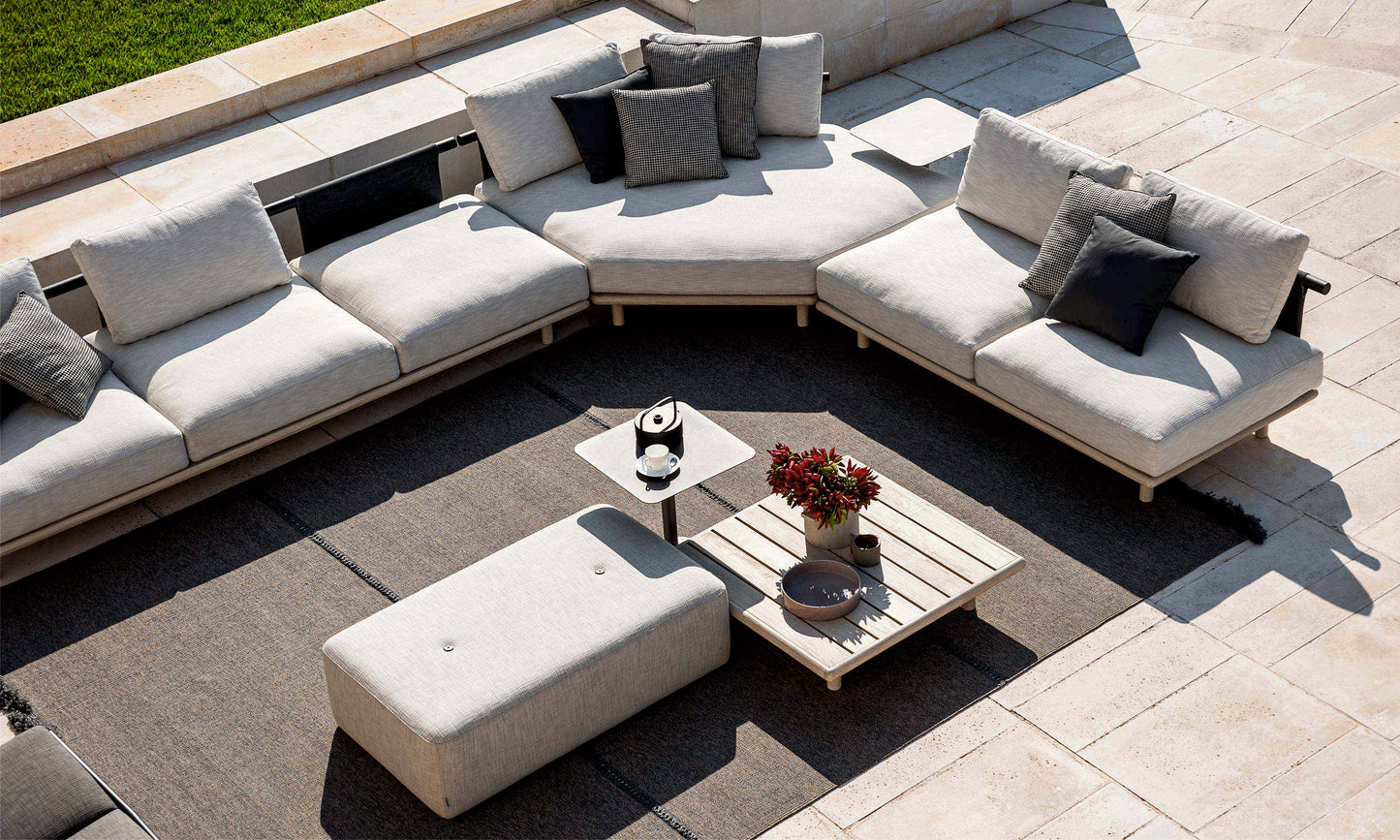 Patio exterior con sofás seccionales de color claro, cojines mixtos, una otomana rectangular y una mesa de centro de exterior SAMBA cuadrada de teca 90 × 90 × 30 cm con un jarrón y tazas sobre una alfombra gris.