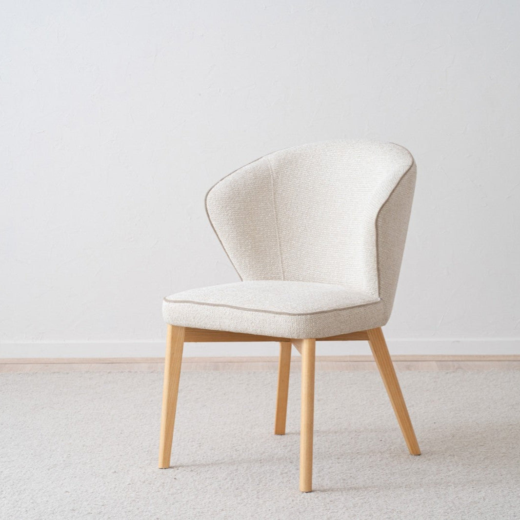 Un moderno Pack 2 Sillas de comedor tapizadas EVA de color beige con respaldos curvados y patas de madera reposa elegantemente sobre un suelo alfombrado claro contra una pared blanca, personificando el diseño contemporáneo.