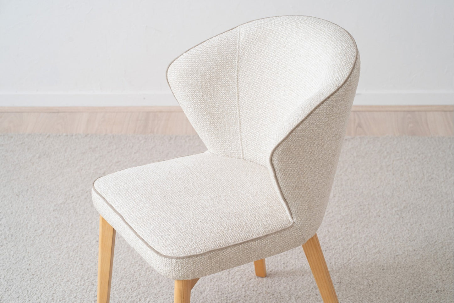 El Pack 2 Sillas de comedor tapizadas EVA, de color crema con patas de madera, se apoya sobre un suelo de moqueta clara, añadiendo un diseño contemporáneo contra la pared blanca lisa.