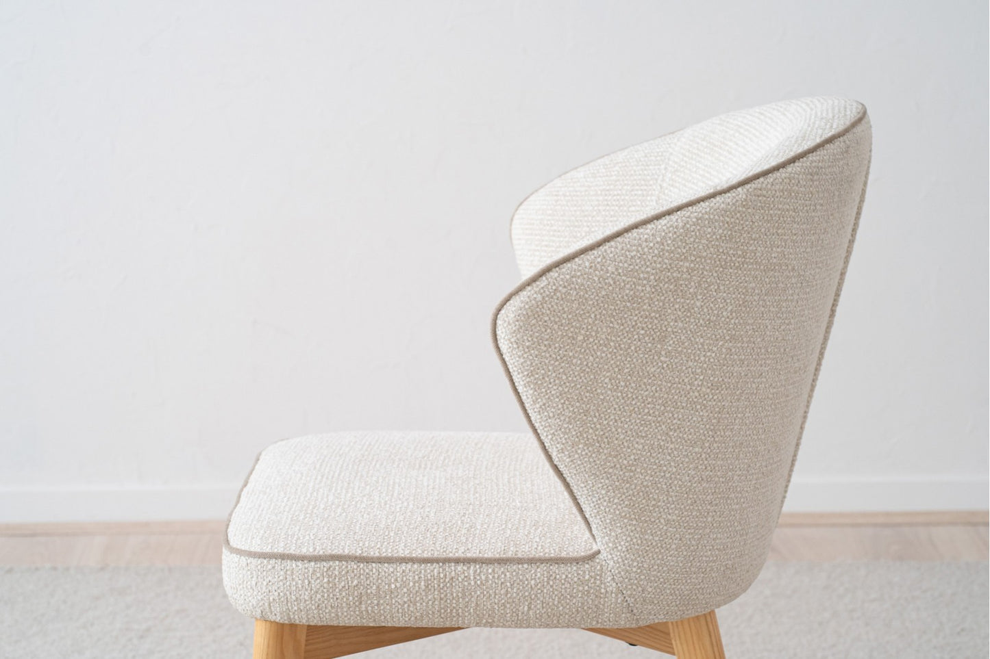 El Pack 2 Sillas de comedor tapizadas EVA, modernas sillas de tela beige claro con marcos de madera, se ven de lado y personifican el diseño contemporáneo contra una pared blanca lisa y un piso de madera clara.