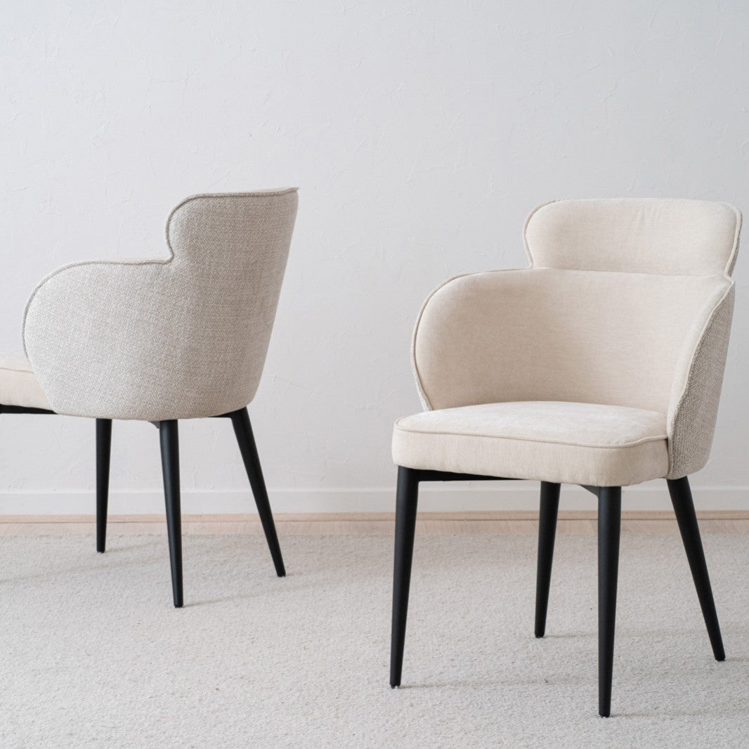 El Pack 2 Sillas de Comedor Tapizadas con Reposabrazos Oslo en color beige presenta un diseño moderno con respaldos curvos y patas negras, elegantemente colocados sobre un piso alfombrado claro contra una pared blanca, mostrando un diseño moderno y funcionalidad.