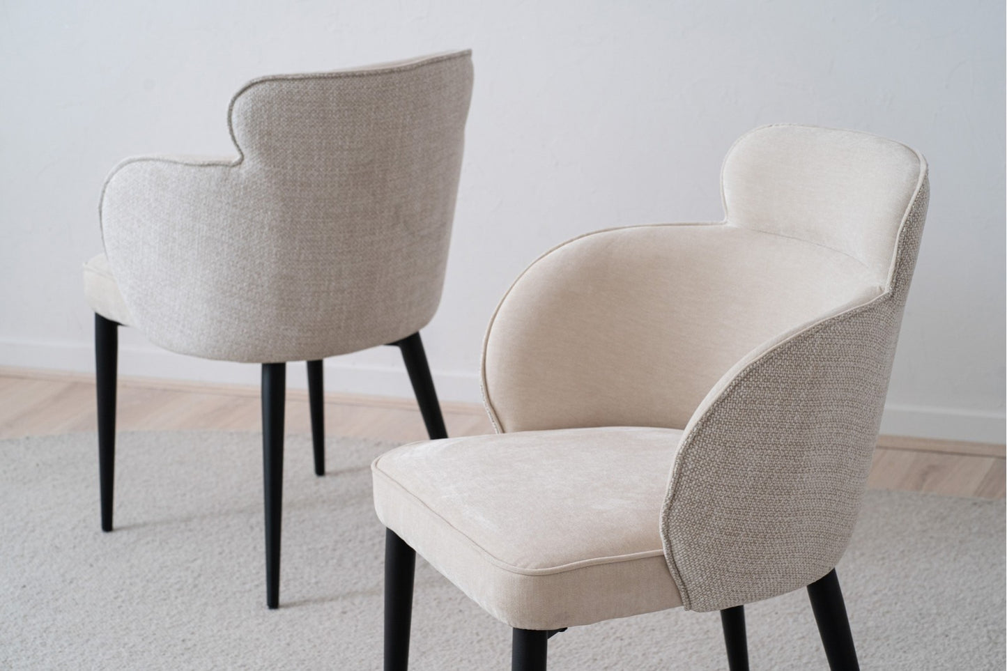 El Pack 2 Sillas de Comedor Tapizadas con Reposabrazos Oslo (Beige) presenta un diseño moderno con tapizado beige y elegantes patas negras sobre una alfombra clara, combinando a la perfección elegancia y funcionalidad en esta habitación minimalista.