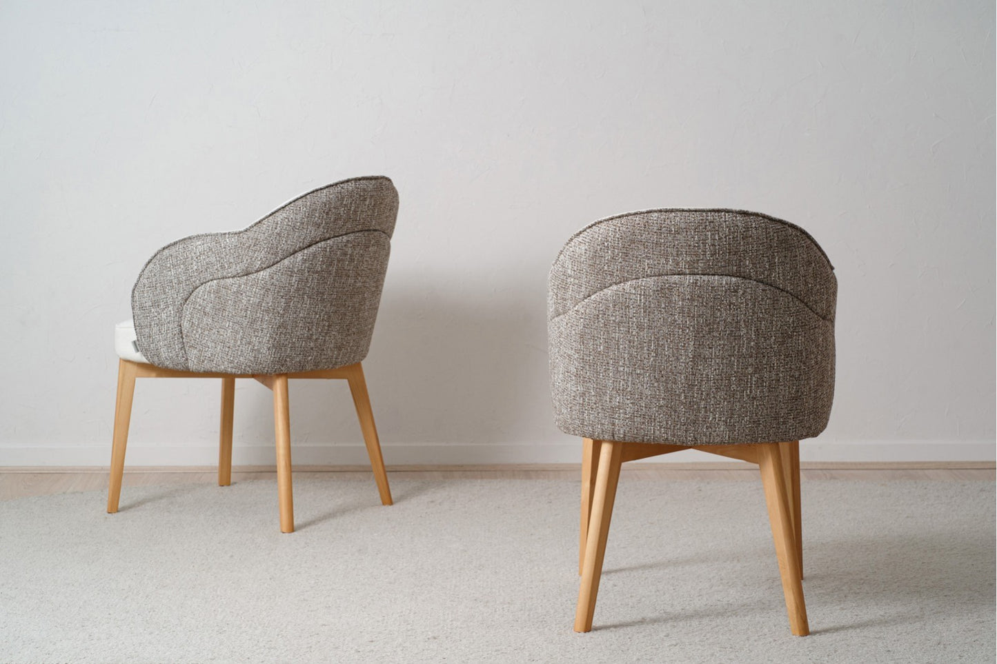 Las Sillas de Comedor tapizadas con reposabrazos Roma Pack 2 en gris, con patas de madera maciza, están elegantemente colocadas sobre una alfombra clara contra una pared blanca nítida, creando un aire de diseño moderno.