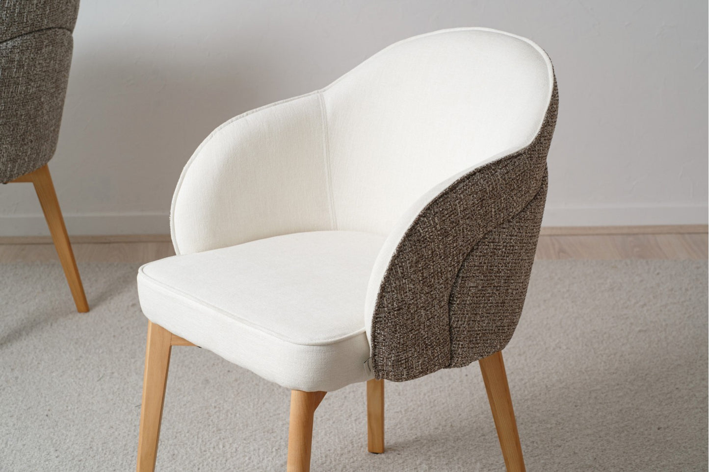 El moderno Pack 2 Sillas de Comedor tapizadas con Reposabrazos Roma (Gris) cuenta con asientos acolchados con tela blanca y gris y patas de madera maciza, elegantemente colocados sobre un piso alfombrado beige.