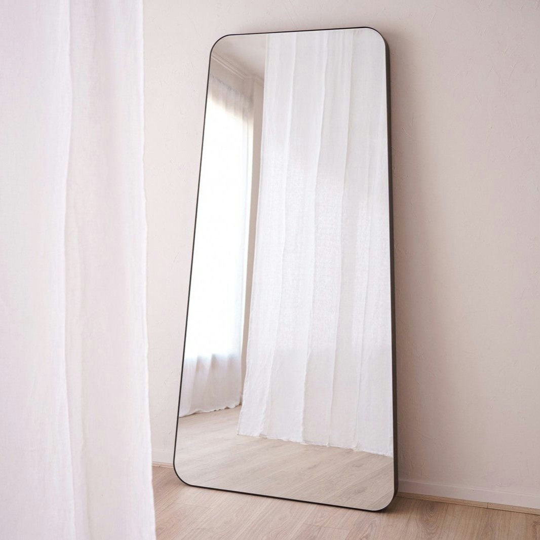 El Espejo de suelo en metal industrial N4, de forma rectangular y esquinas redondeadas con marco metálico, se apoya contra una pared blanca en una habitación con cortinas blancas translúcidas y suelo de madera clara.